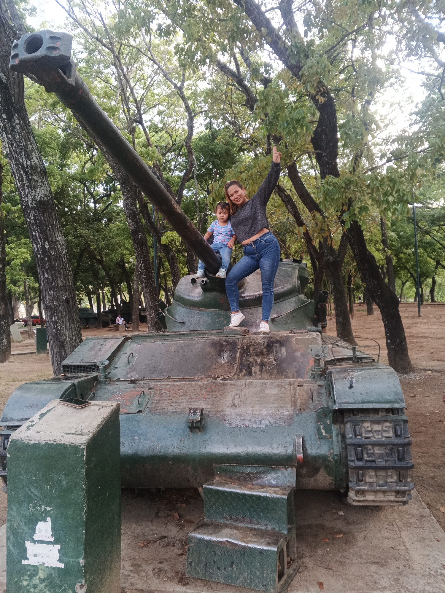 A woman and a young child are standing on a green, decommissioned military tank displayed in a park. The woman, wearing a gray shirt and blue jeans, stands on the tank with one leg bent and her arm raised in a peace sign. The child, in a blue shirt and jeans, sits atop the barrel of the tank. The tank is positioned in a park setting with tall trees in the background. A concrete block with a mobile phone on top serves as a step to mount the vehicle. The location is Caracas, Venezuela.