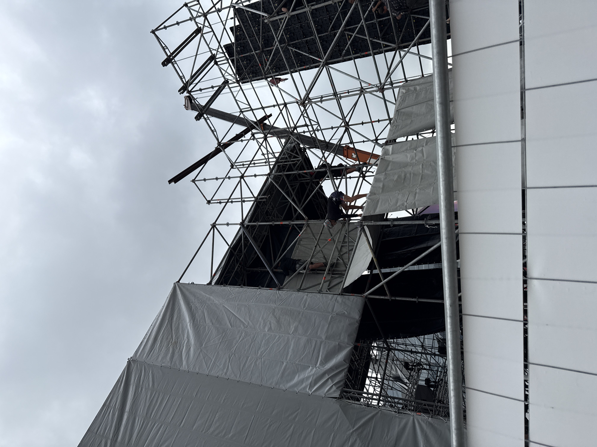 A medium shot captures a large outdoor stage construction under a cloudy sky. Scaffolding structures dominate the frame, covered in large white tarpaulins on the left, obscuring parts of the stage. The scaffolding itself is a complex network of metal poles and supports, with some sections covered by dark fabric. A crane arm is visible extending from the top right, its orange boom partially obscuring the metal framework. Several figures are engaged in work on the scaffolding, dressed in casual clothing. One person is seen leaning over a platform, another is climbing, and a third is partially visible near the bottom right. In the foreground, a white mesh barrier with a metal top rail separates the viewer from the construction site, suggesting public access is restricted. The overall scene depicts an ongoing construction or dismantling process for a large event. The location is not discernible from the image itself.