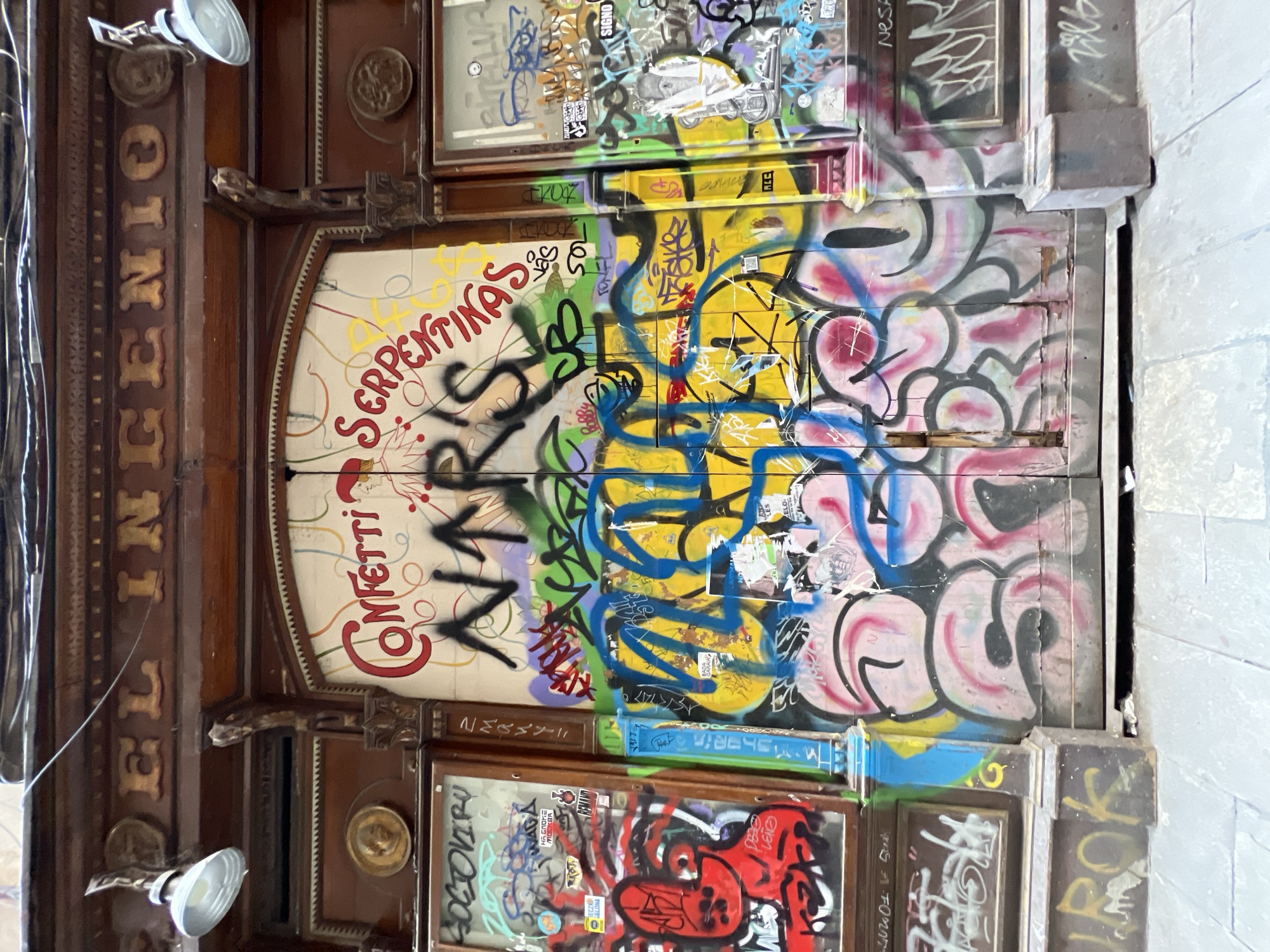 Here is a description of the image:
The image depicts an ornate wooden door, heavily covered in graffiti, in what appears to be Barcelona, Spain. The door has a central arched panel with the words "Confetti" and "Serpentinas." The word "INGENIO" is vertically written in gold letters on the left side of the door frame. Two glass-paneled sections are above and below the central panel. All surfaces of the door and panels are extensively tagged and layered with colorful graffiti. The surrounding wall is plain and faded. The door and its framing are weathered and show signs of age. There are two spotlights on the top of the left side of the door.