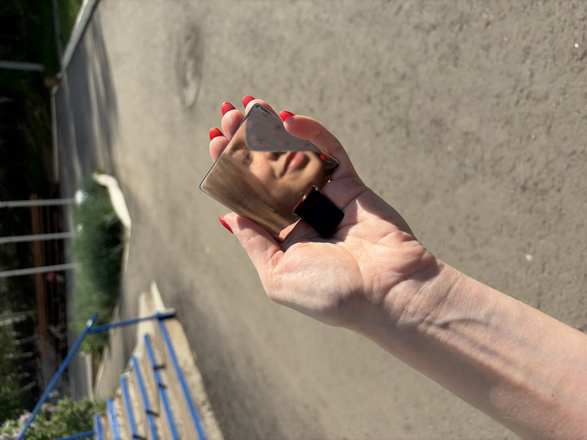 A woman's arm and hand are outstretched, holding a small, reflective object. The object seems to be a perfume bottle, reflecting the woman's face within its surface. She has red nail polish on her fingernails. The background suggests an outdoor setting, specifically a paved area, possibly a road or walkway. Parts of a blue railing and green vegetation are visible in the peripheral view, along with sections of a building. This scene is located in the city of Chaikovsky, Russia.