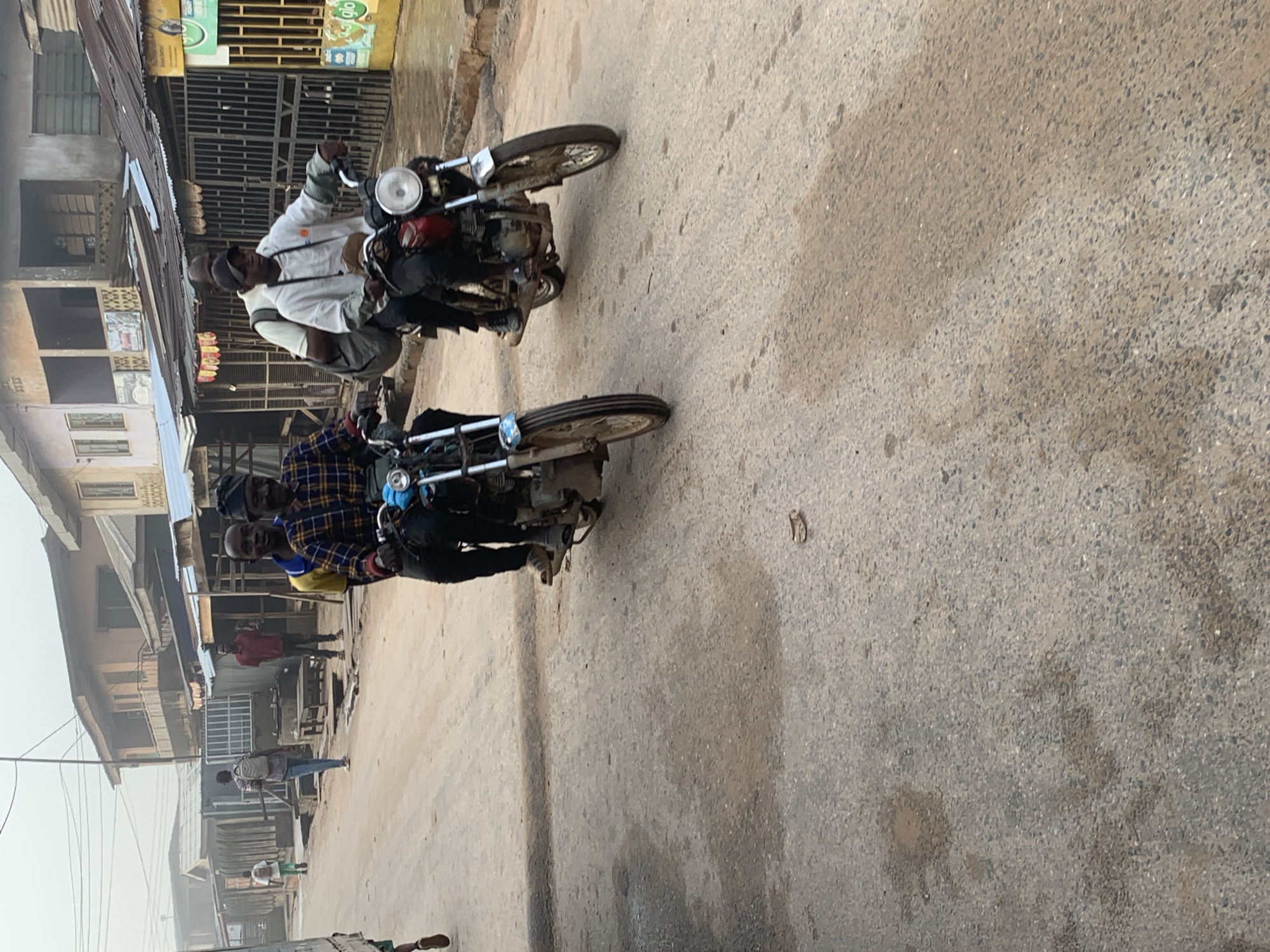 The image depicts a street scene in Shagamu, Nigeria, characterized by dusty, uneven asphalt pavement and buildings with corrugated metal roofs lining both sides of the road. Two motorcycles are prominently featured. The first motorcycle, closer to the foreground, carries two individuals. The rider wears a blue and yellow plaid shirt and a black cap. The passenger behind him wears a yellow shirt and a blue collar. The second motorcycle, further down the road and slightly to the right, is ridden by a person wearing a white hooded sweatshirt and a black baseball cap. This motorcycle also carries a passenger wearing a white t-shirt and a backpack. Other individuals are visible in the background, including a person in a red shirt and another carrying a backpack. The environment suggests a commercial or residential area with a somewhat unkempt appearance, common in many Nigerian towns. The sky is overcast, contributing to a muted light.
