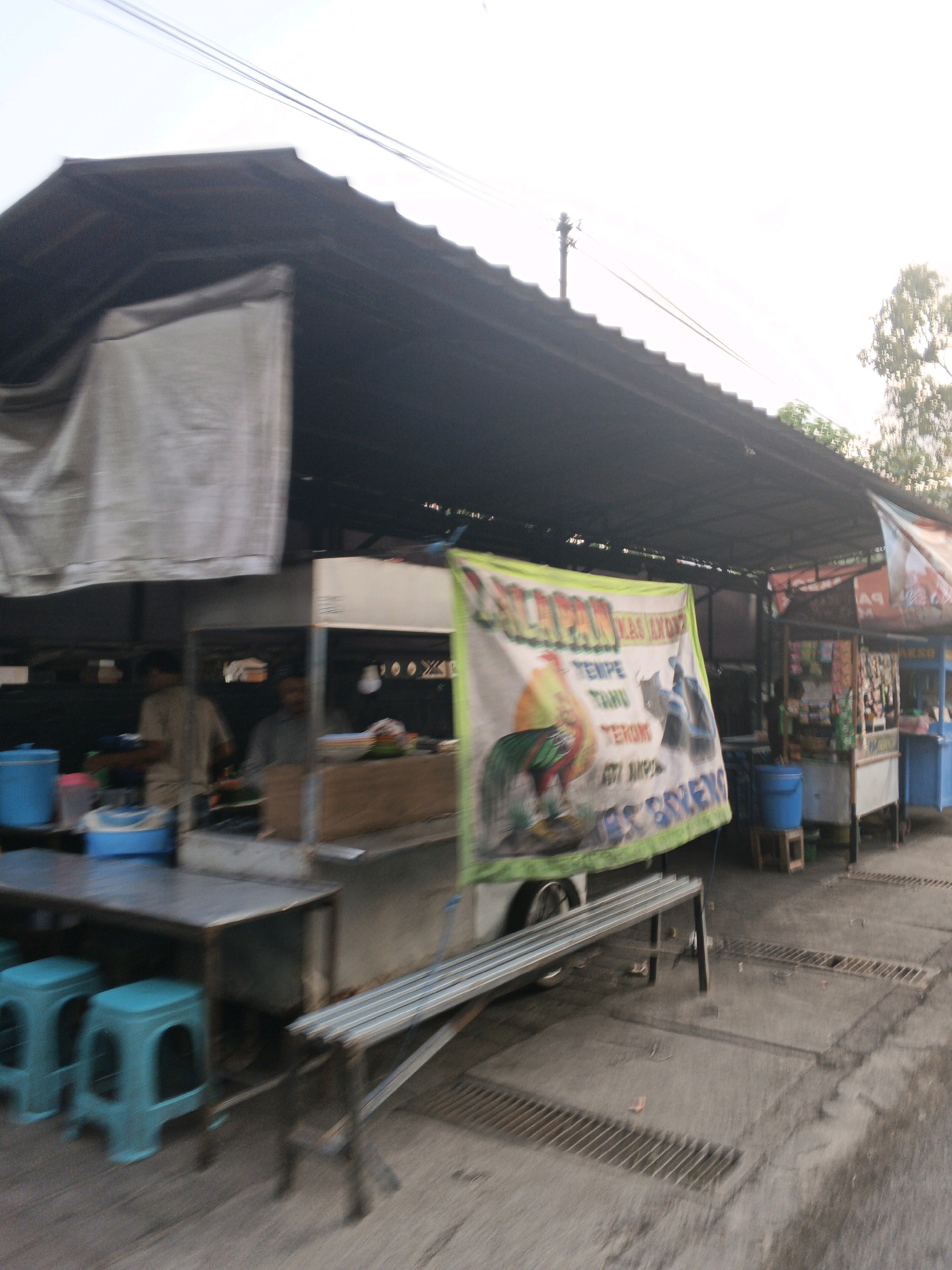The image shows an outdoor food stall in Sukawati, Indonesia. The stall has a dark, sloped roof extending over a covered area. A large banner with text and a colorful rooster graphic is prominently displayed on the front of the stall. To the left, there are blue plastic stools and a table. In the background, there are more stalls with merchandise and food items. Two individuals, appearing to be vendors, are visible behind the counter area of the stall. The ground is paved and features drainage grates. The overall scene depicts a bustling local market environment.