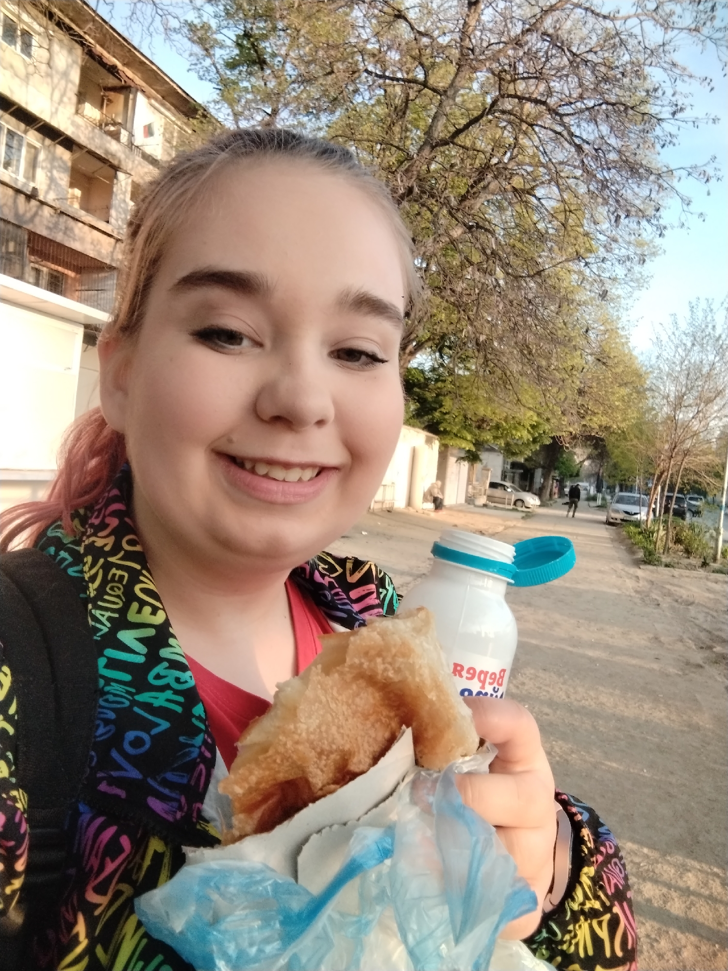 A woman holds food in a wrapper and a bottle on a street in Plovdiv, Bulgaria. The woman is facing the camera and smiling. She has light skin, pink hair, and is wearing a black jacket with colorful letters and a red shirt. She is holding food in a white wrapper and a plastic bag in her right hand. In her left hand, she is holding a white bottle with a blue lid. The street has a dirt path, and there are trees lining the street. Cars are parked on the side of the street, and a person is walking in the distance. A building can be seen in the background.