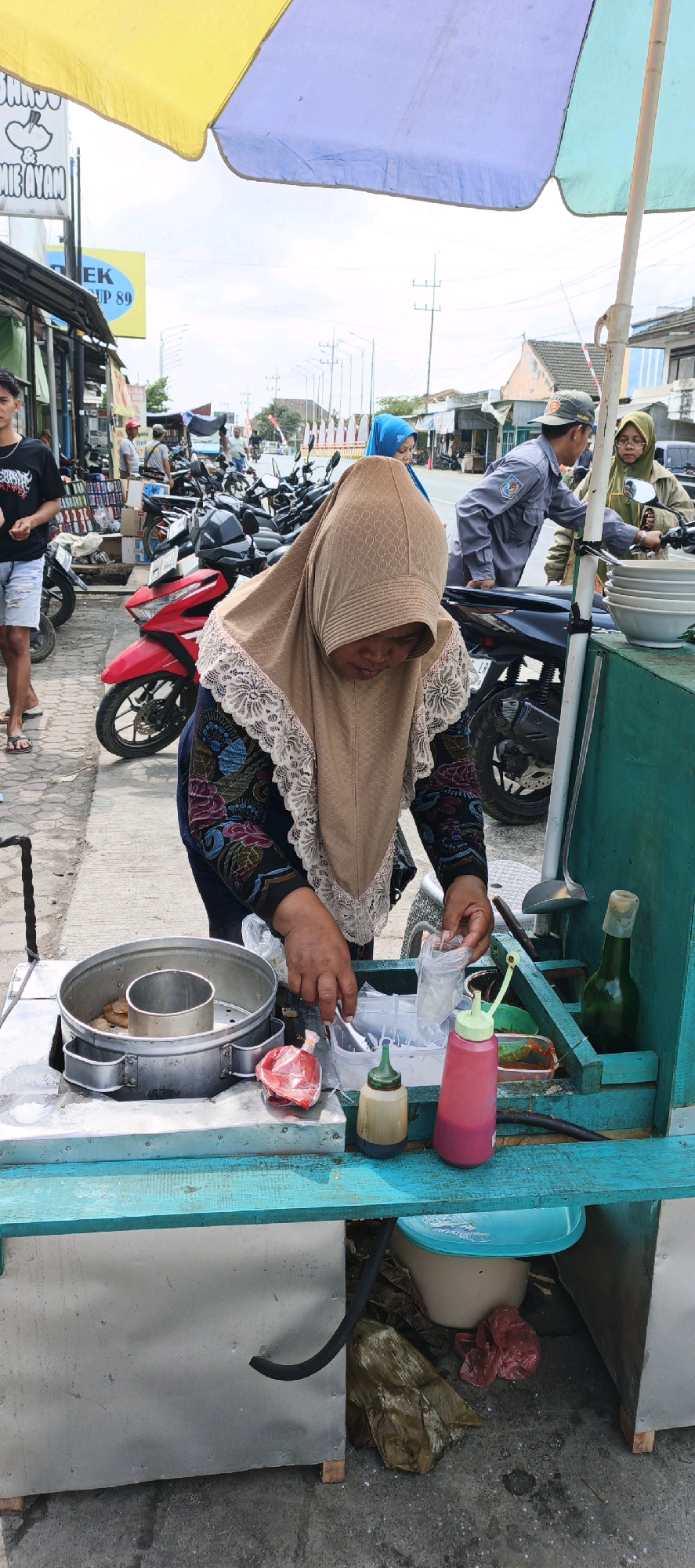 In Kasiyan, Indonesia, an outdoor food stall operates under a colorful umbrella. A woman wearing a tan hijab and a patterned blouse is actively preparing food. Her hands are engaged in packaging items, suggesting she is serving a customer or preparing for one. In front of her, a metal steamer contains food, and various containers of condiments and utensils are arranged on the teal-colored counter of the mobile cart. The cart itself appears to be made of metal and wood, with a black hose attached to its base. Behind the stall, a street scene unfolds with parked motorcycles, other stalls, and pedestrians. The sky is overcast.