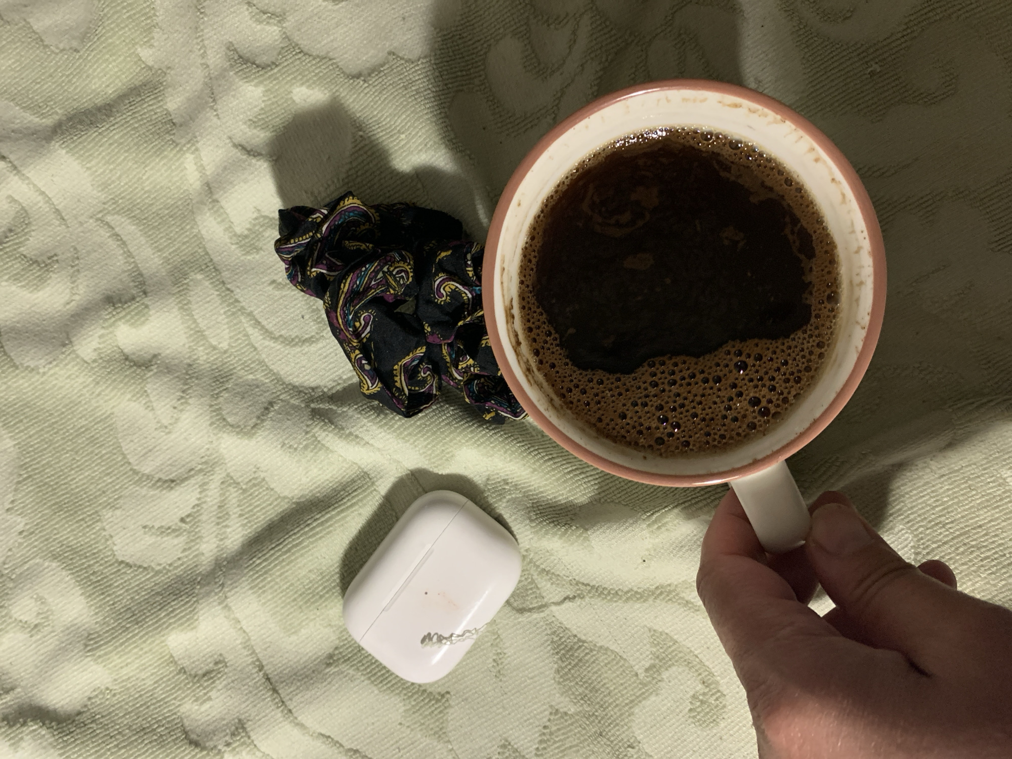 An overhead shot shows a light green textured blanket. On the blanket, to the left of center, is a white earbud case. To the right of the earbud case is a black scrunchie with a paisley pattern in purple, yellow, and teal. In the lower right quadrant of the image, a hand is holding a pink and white mug filled with dark coffee, which has a foamy surface. The mug is positioned to the right of the scrunchie and earbud case.