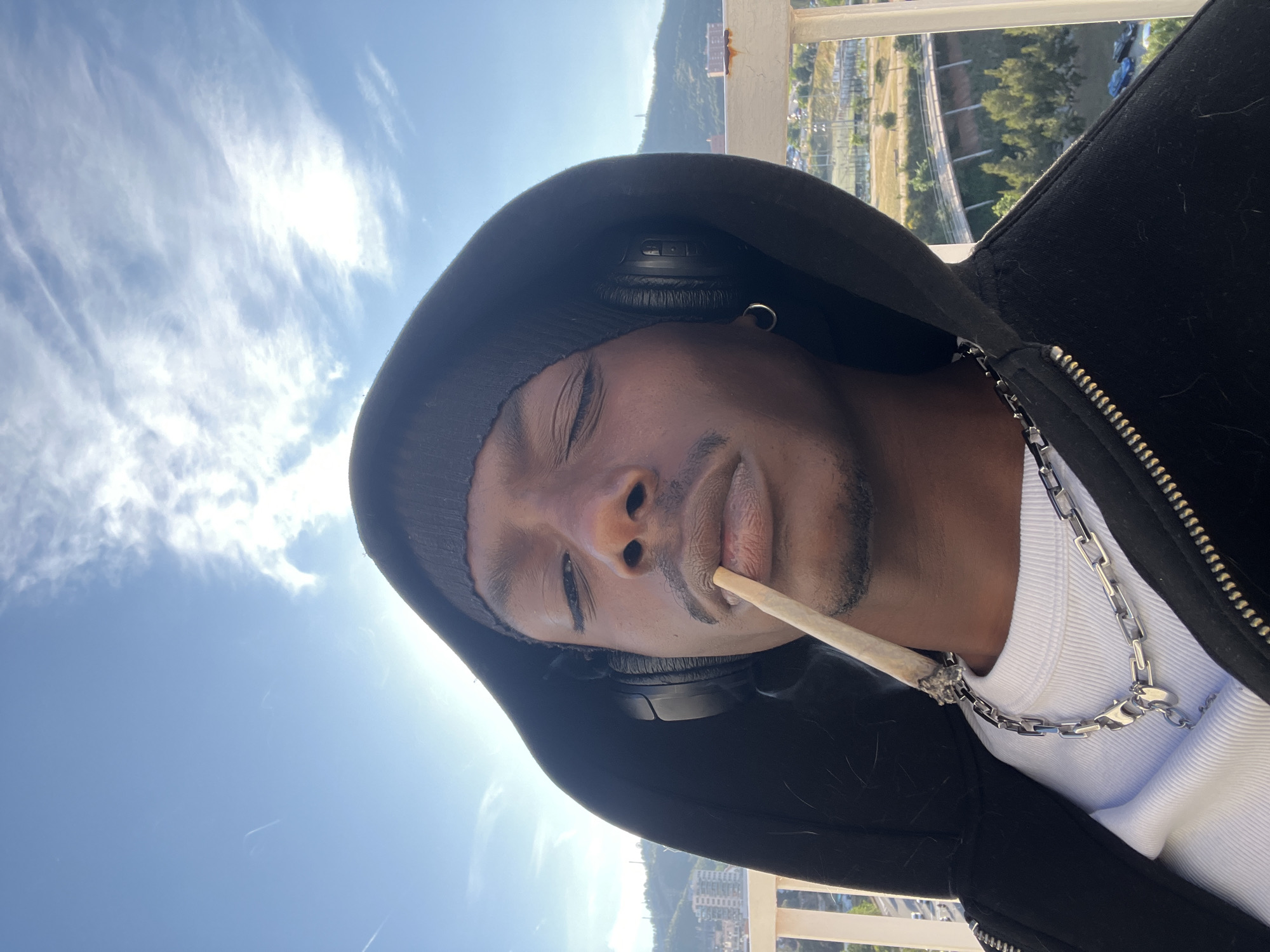 A medium shot captures a Black man with his eyes partially closed, exhaling a rolled cigarette. He is wearing a black hooded jacket over a white ribbed tank top, adorned with a silver chain. Headphones are positioned around his ears, partially covered by a black beanie. An earring is visible on his left ear.
The backdrop showcases an urban landscape featuring buildings and trees, with a mountainous region in the distance. The scene is bathed in natural light under a sky with light clouds. The location appears to be a rooftop balcony or elevated platform. While I can not definitively ascertain the exact location in Barcelona, the mountainous region in the distance suggests that the image was taken overlooking the city from an elevated platform in the Collserola hills.