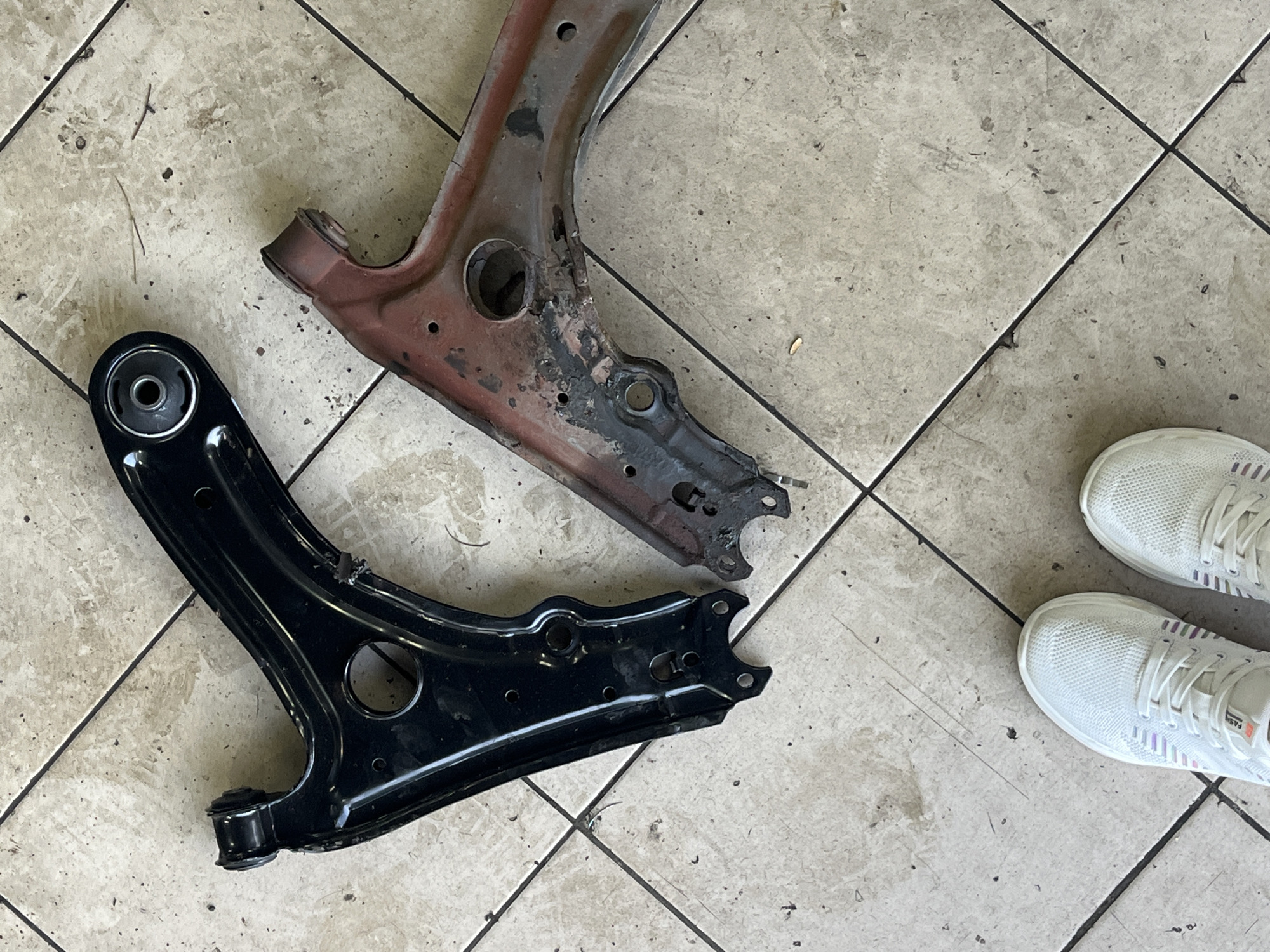 Two used car control arms, one black and one red, are laid out on a tiled floor. The black control arm is on the left, and the red one is on the right. Both appear to be for the front suspension of a vehicle. The red control arm shows significant rust and damage, suggesting it is a discarded part. In the lower left corner of the image, a pair of white athletic shoes are visible. The environment appears to be an indoor space, possibly a garage or workshop, with a light-colored, square tiled floor. The image is taken from a top-down perspective. The location is not specified in the image.
