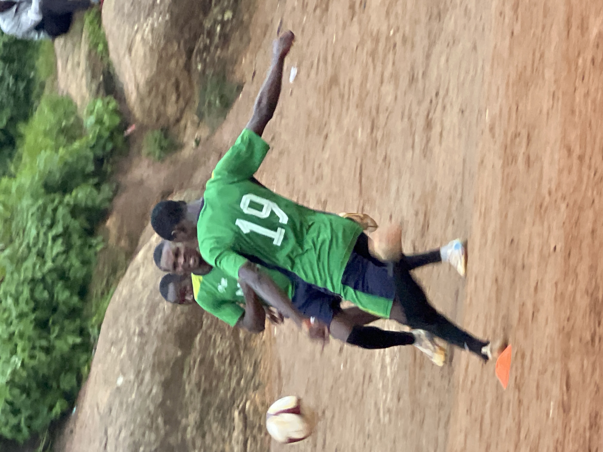The image depicts an outdoor soccer training session in Jos, Nigeria. The ground is a dirt or sand surface, common in many areas of the city. In the foreground, a young man wearing a green soccer jersey with the number "19" on the back is in motion, appearing to kick a soccer ball that is blurred due to movement. He is also wearing dark shorts and long black socks. To his left and slightly behind him, another young man in a similar green jersey is also in motion, possibly engaged in the same drill or play. Further behind them, a third individual in a green jersey is visible. In the background, there are large rocks and lush green vegetation, typical of the natural landscape surrounding Jos. The lighting suggests it is daytime, likely overcast or late afternoon given the soft light. The overall scene captures a moment of athletic activity in a natural, somewhat unmanicured environment.
