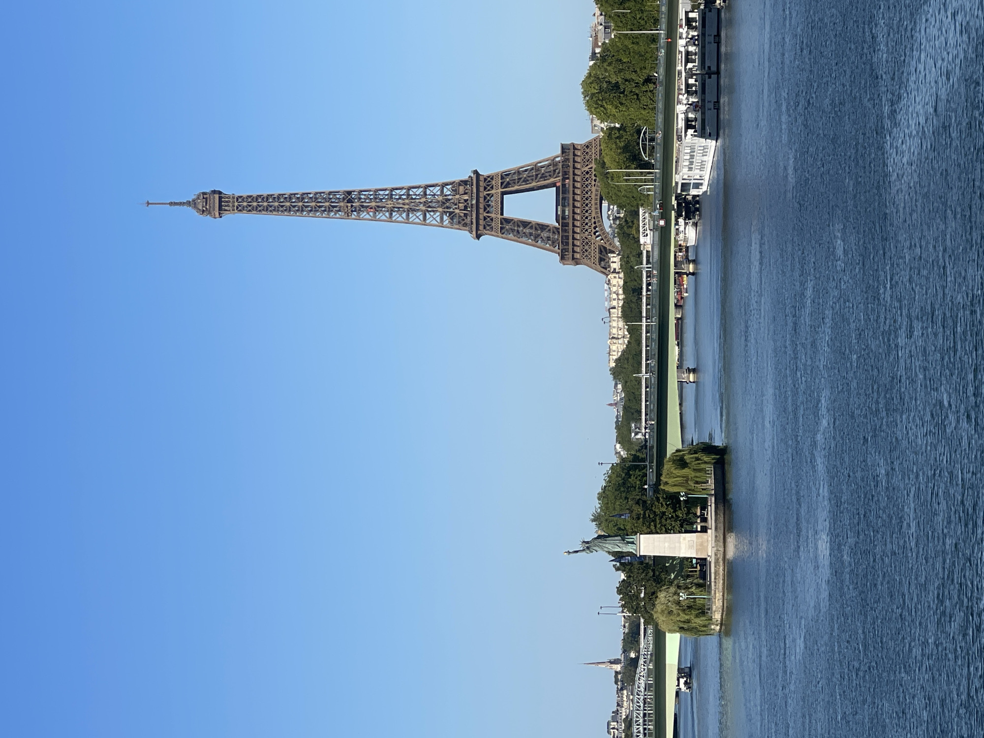 The image shows a view of the River Seine in Paris, France, on a clear day with a bright blue sky. In the foreground, the rippling blue water of the Seine occupies the lower half of the frame. In the middle ground, a small island with trees and a statue of Liberty is visible. The statue, a replica of the Statue of Liberty, stands on a pedestal, facing to the right. It is surrounded by lush green trees, with weeping willows on either side. A lamppost is positioned on either side of the statue.
Dominating the background is the iconic Eiffel Tower, a tall wrought-iron lattice tower, its structure clearly defined against the sky. To the left of the Eiffel Tower, a distant cityscape of Paris can be seen, featuring buildings and a spire. Several bridges cross the Seine, with the closest one being a green-painted bridge with visible railings. Boats, including what appear to be tour boats, are moored along the riverbanks or navigating the water. The overall scene captures a serene moment in Paris, showcasing its famous landmarks and the tranquil flow of the Seine River.