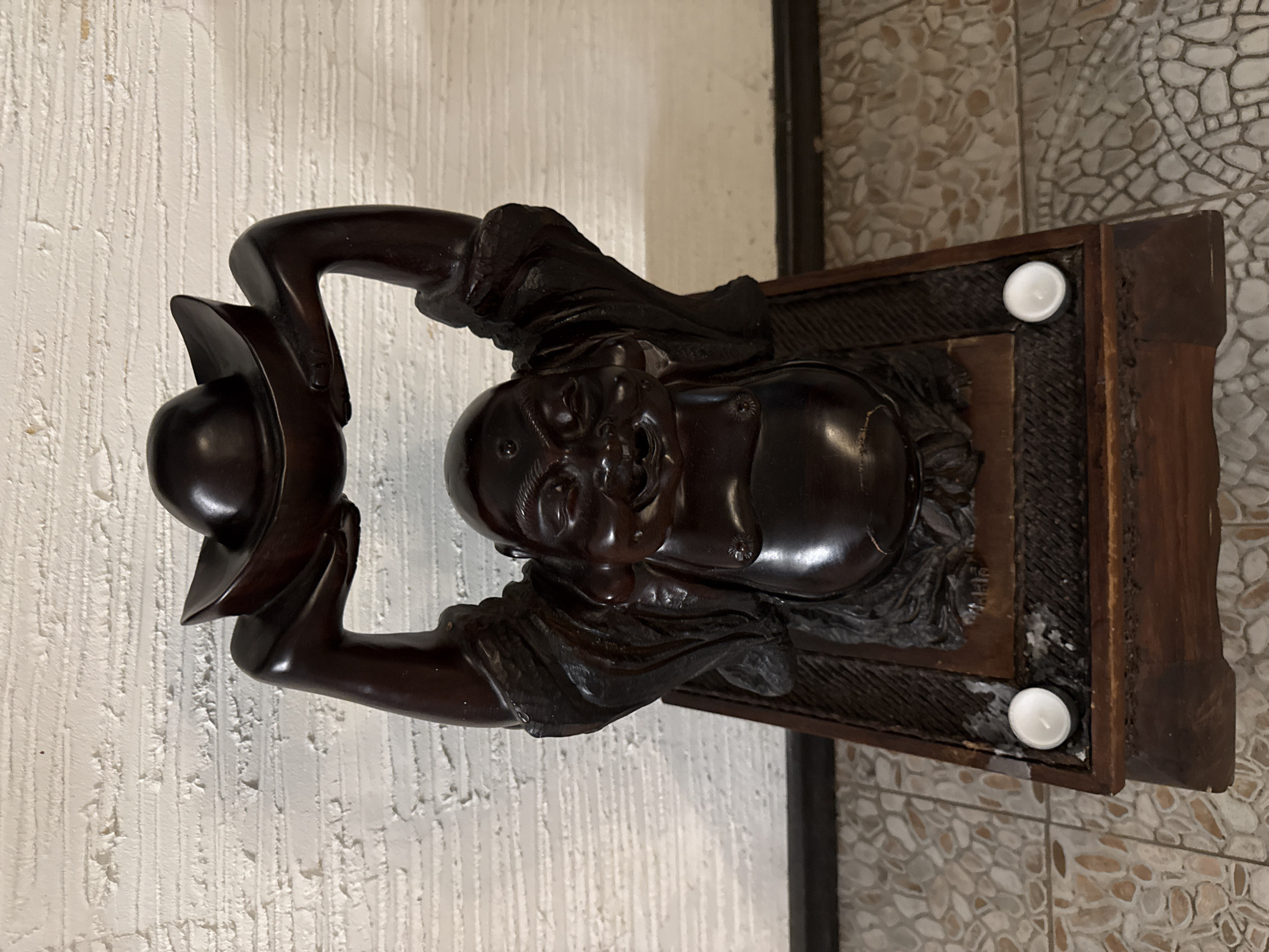 A dark wood carving of a smiling Buddha figure, with arms raised above its head holding a curved, bowl-like object, is placed on a dark wood stand. The stand has intricate carvings and two small white votive candles placed on its surface. The figure and stand are positioned in front of a textured, off-white wall with visible horizontal lines. The floor beneath the stand is tiled with a mosaic pattern of light and dark colored stones. The scene is illuminated, suggesting indoor lighting. There are no people or animals present. The location is identified as Bansko, Bulgaria.