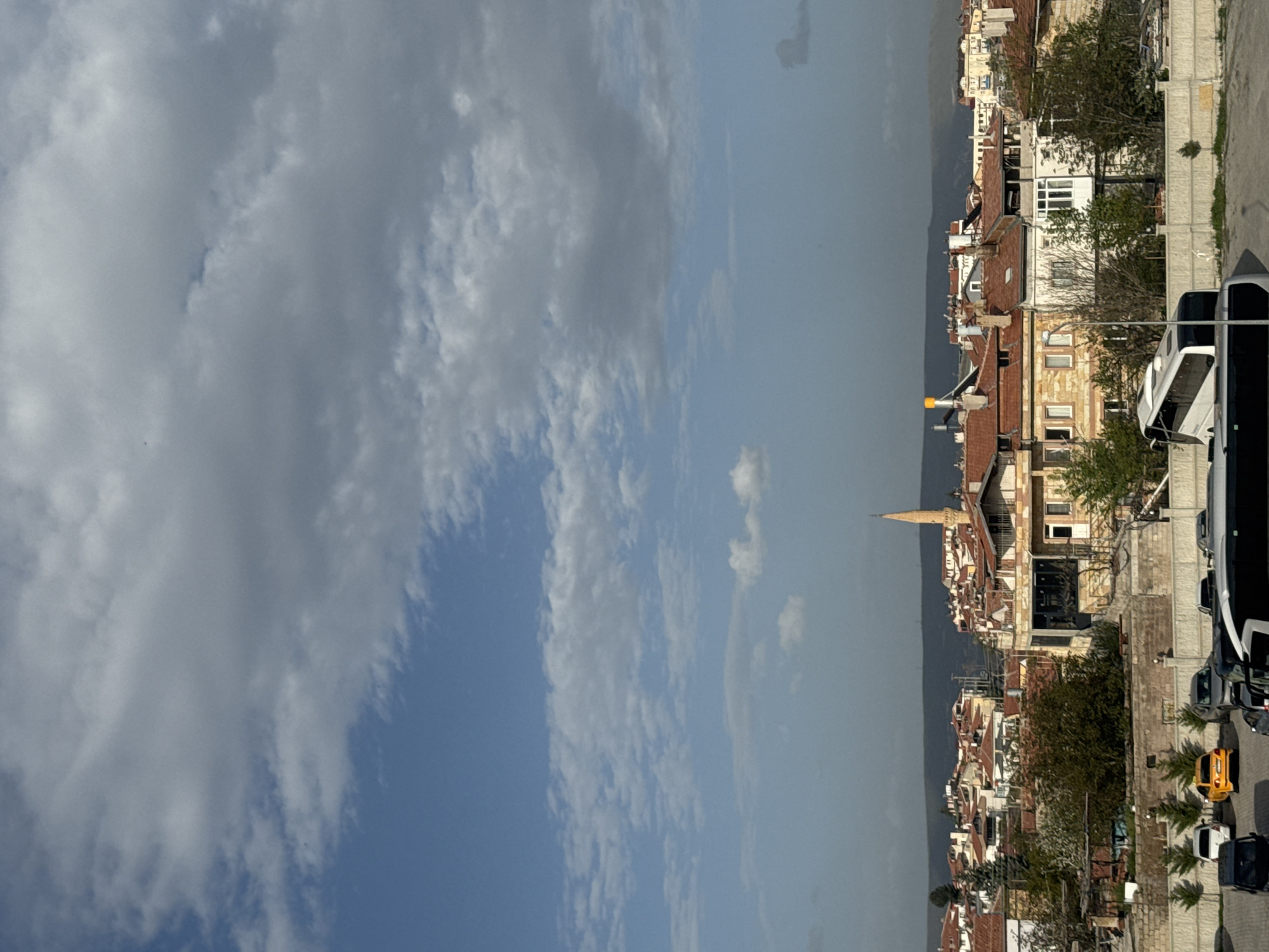 The image shows a view of Ürgüp, Turkey. The right side of the image captures a street scene with buildings, trees, vehicles, and a sidewalk. The buildings are various heights and styles with red-tiled roofs. Vehicles are parked along the street, including a yellow car and a large bus. The left side of the image is filled with a sky with cloudy skies. A thin mountain silhouette is in the center of the image, separating the sky and the buildings. The minaret of the mosque dominates the horizon line.