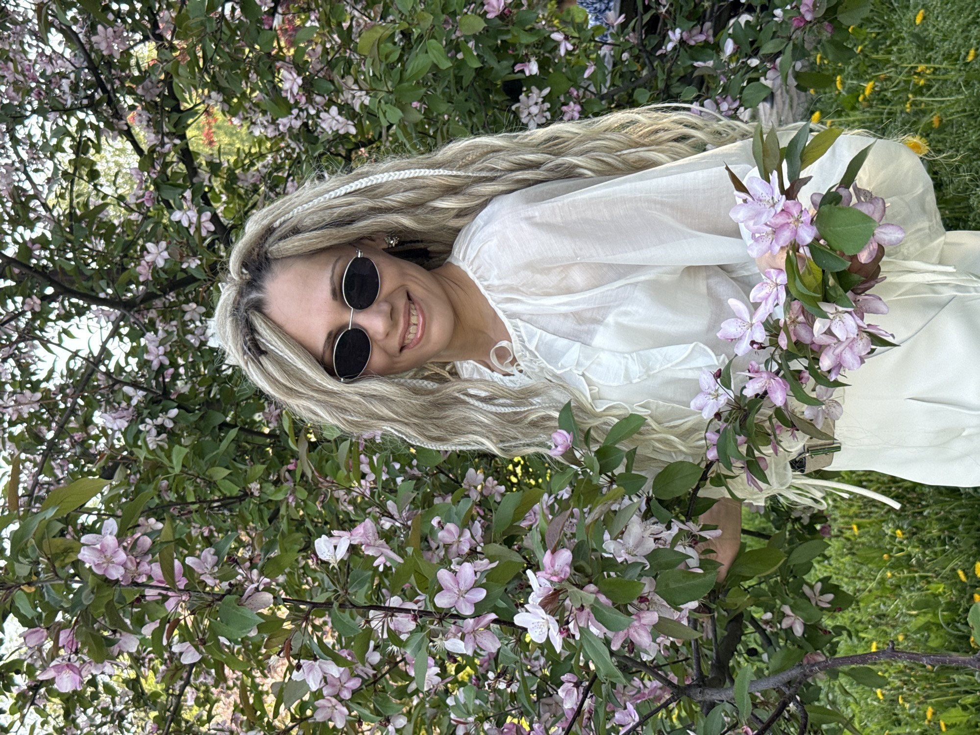 A smiling woman with long blonde dreadlocks and sunglasses is posing outdoors, likely in a park or garden setting. She is wearing a white blouse and what appears to be a matching dress or pants. She is surrounded by flowering trees with pink and white blossoms and green foliage. The background also includes green grass. The location is suspected to be Rusanovo, Russia.