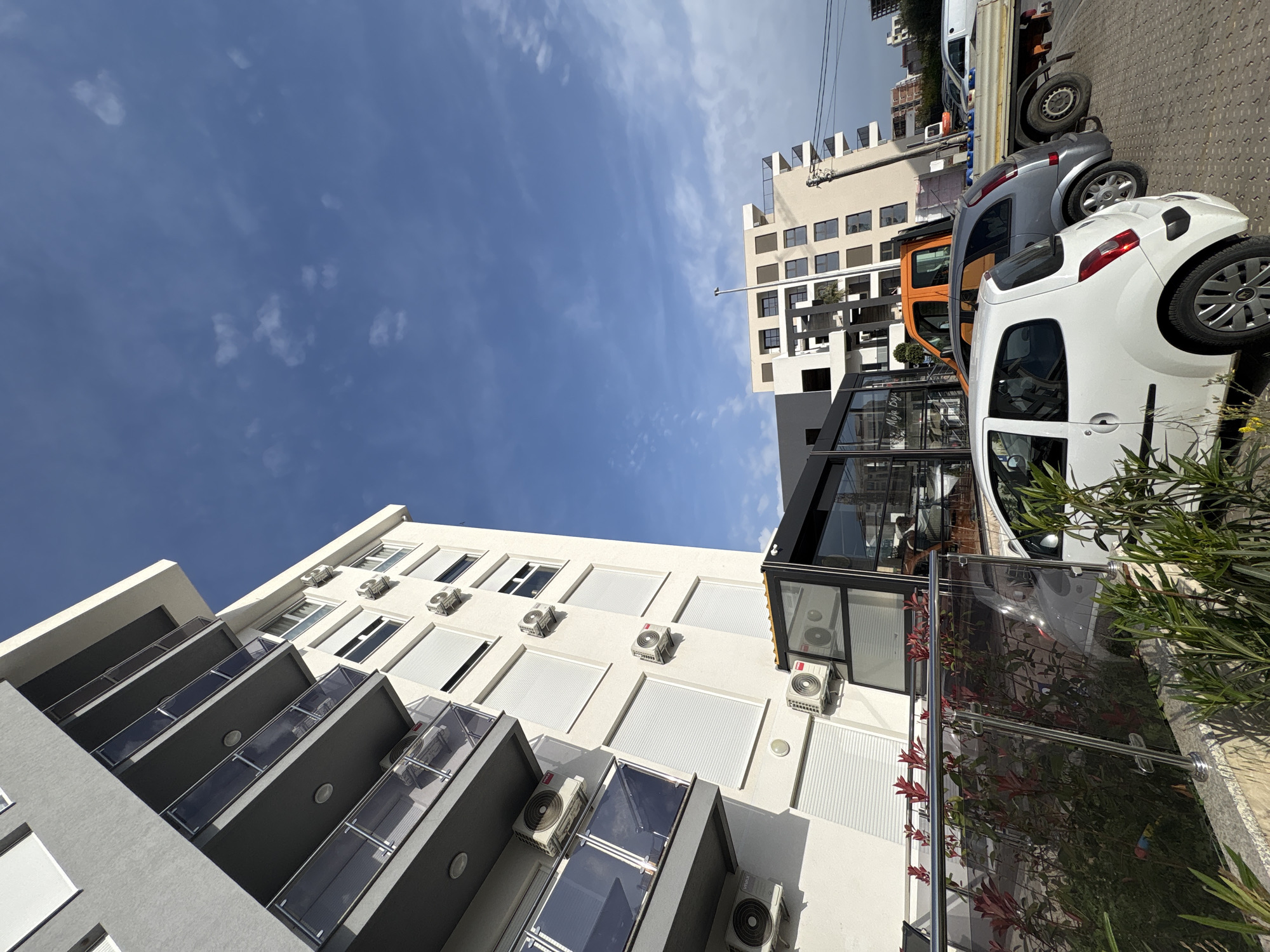 The image shows an outdoor scene in Bar, Montenegro. It is taken from a low-angle perspective, looking up at a building with white walls, dark grey balconies, and numerous air conditioning units mounted on the facade. Several parked cars are visible along the side of the building, including white, grey, and orange vehicles. Another building with multiple windows is seen in the background. Above the buildings, a blue sky with scattered clouds is visible. Plants and some reflective surfaces also appear in the scene.