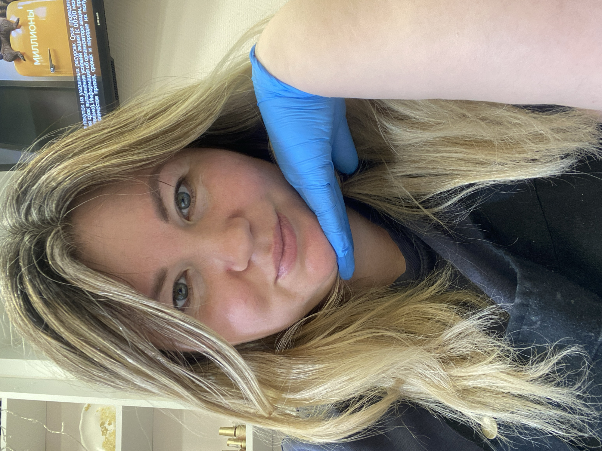 The image captures a close-up portrait of a woman with long, wavy blonde hair, light skin, and blue-green eyes, looking directly at the camera with a calm, slightly smiling expression. Her head is tilted, and her hair falls around her shoulders and face.
Her right arm and hand, clad in a bright blue nitrile or latex glove, are visible, with the gloved hand resting gently against her jawline and cheek. This detail suggests she might be undergoing or has just completed a procedure, possibly related to beauty, dental work, or a medical examination, or perhaps she is performing such a procedure herself.
In the background, a television screen is visible, displaying text in Russian. The word "МИЛЛИОНЫ" (MILLIONS) is prominent, along with other text describing "покупок на указанных ресурсах. Срок проведен... выполнения условий акции... организаторе акции, призов, сроках и порядке их полу" (purchases on the specified resources. Period of... fulfillment of action conditions... organizer of the action, prizes, terms and conditions of their receipt). The brand "SAMSUNG" is also visible on the TV. Below the TV and to the woman's left, parts of white shelves containing what appear to be decorative items can be seen.
The setting appears to be indoors, possibly in a salon, clinic, or a well-lit room in a private residence in Chaikovsky, Russia. The lighting is bright and even, indicating daytime or good artificial illumination.