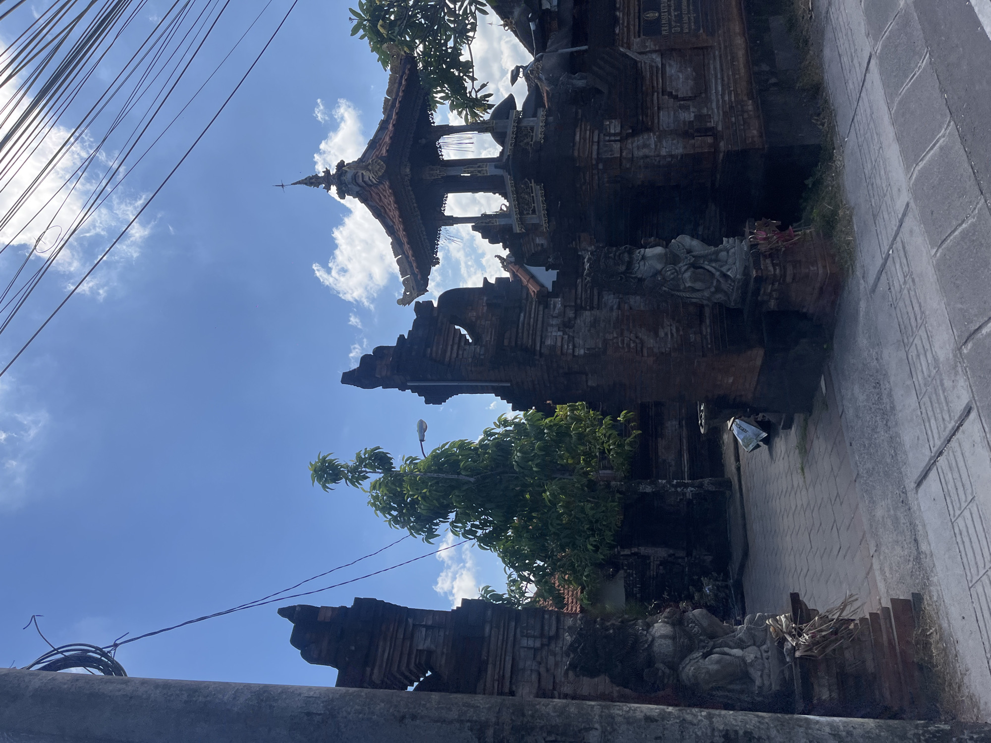 A low-angle, medium shot captures a traditional Balinese temple entrance with weathered brick structures and ornate carvings under a bright blue sky dotted with scattered clouds. The gate features two imposing stone statues of guardians, one on each side, at the entrance. A lush green tree stands tall between the gateway pillars. Overhead, a network of dark electrical wires crisscrosses the sky, suggesting an urban or semi-urban setting within Denpasar Barat, Indonesia. The paved walkway leads into the temple courtyard, where a tiered, pavilion-like structure with a red tile roof is visible in the background. A street sign is also partially visible on the right side of the image.