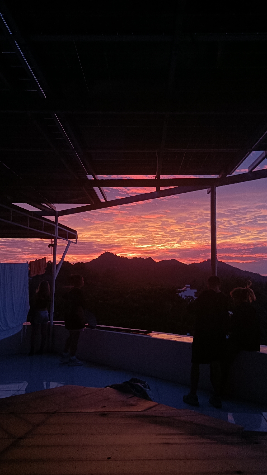 The image depicts a sunset view from a rooftop balcony in Nha Trang, Vietnam. Silhouetted figures of at least five people are visible, gathered on the balcony, facing away from the camera and towards the horizon. The sky is filled with vibrant colors of orange, pink, and purple, indicative of a dramatic sunset. In the background, dark, rolling hills or mountains are visible, with some hints of buildings on one of the slopes. The foreground shows part of the balcony structure and a table or platform covered with a reddish-brown cloth. Laundry is hanging on a line to the left of the frame. The overall atmosphere is serene and contemplative, with the people appearing to be enjoying the scenic view.