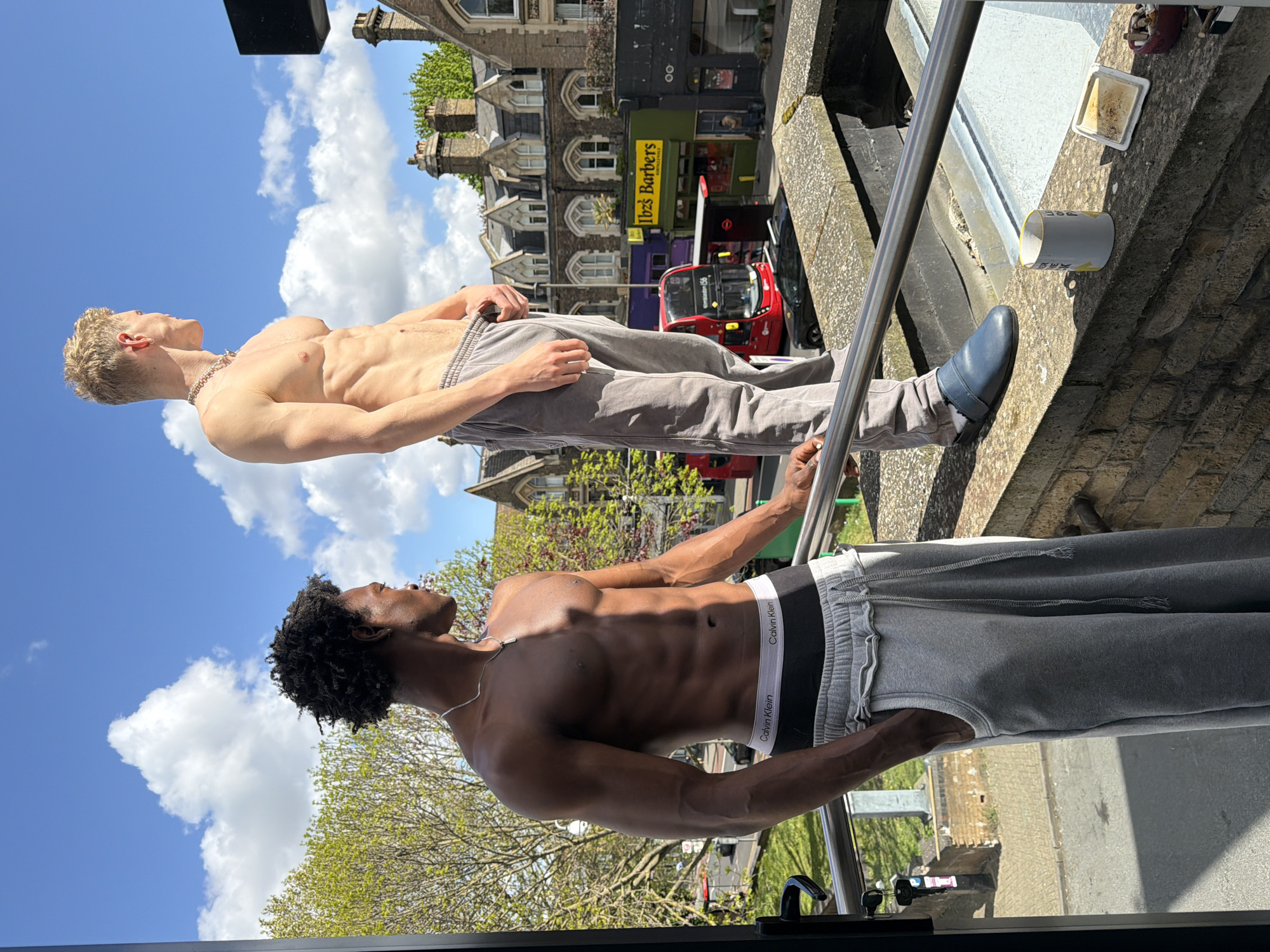 The image shows two shirtless, athletic men on a balcony overlooking a street in London, United Kingdom. One man has dark skin and curly black hair, wearing grey Calvin Klein underwear and grey sweatpants. The other man has pale skin and blonde hair, wearing grey sweatpants and blue shoes. Both men are looking up. The background includes buildings, a red double-decker bus, a black car, a shop named "Ibiza Barbers" and a partly sunny sky.