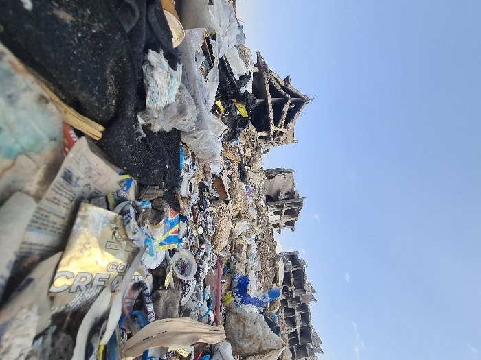 The image depicts a static, expansive scene within Joubar Municipality, Syria, dominated by accumulated refuse and structural devastation under a clear, light blue sky.
The foreground is occupied by a dense, heterogeneous layer of discarded materials. This includes prominent black textile or fabric, several soiled disposable diapers with blue patterns, various plastic packaging materials such as a blue and yellow wrapper and transparent plastic sheeting, fragments of newspaper and other paper waste, a crumpled gold-colored foil wrapper with partial text "CREA" visible, a white disposable plate, and a wooden splinter. This refuse forms an uneven terrain extending into the midground, mixed with general rubble and dirt.
The background features three severely damaged, multi-story buildings silhouetted against the sky. The structure on the far left presents a distinct, partially collapsed roofline with a triangular or gabled appearance, exposing multiple internal floor levels and structural elements. The central and right-most buildings also exhibit extensive structural damage, characterized by missing wall sections, exposed concrete slabs, and skeletal frameworks, indicative of prolonged degradation or severe impact.
No individuals, actions, or direct interactions are visible within the frame. The environment suggests an abandoned urban area significantly impacted by conflict or disaster, now serving as a site for waste accumulation.