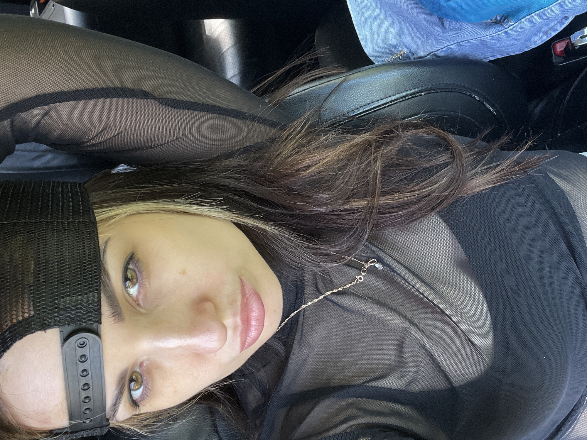A young woman lies on a black leather seat, likely inside a car, as inferred from the surrounding dark interior and a fragment of a denim garment visible in the upper right corner. She is wearing a black mesh top over what appears to be a black bra, and black tights that are also a mesh or net-like material. A black baseball cap sits on her head. A thin silver necklace adorns her neck. Her hair is long and brown with blonde highlights. Her eyes are open and she's looking directly at the camera with a neutral expression. The location of the photo is Unknown.
