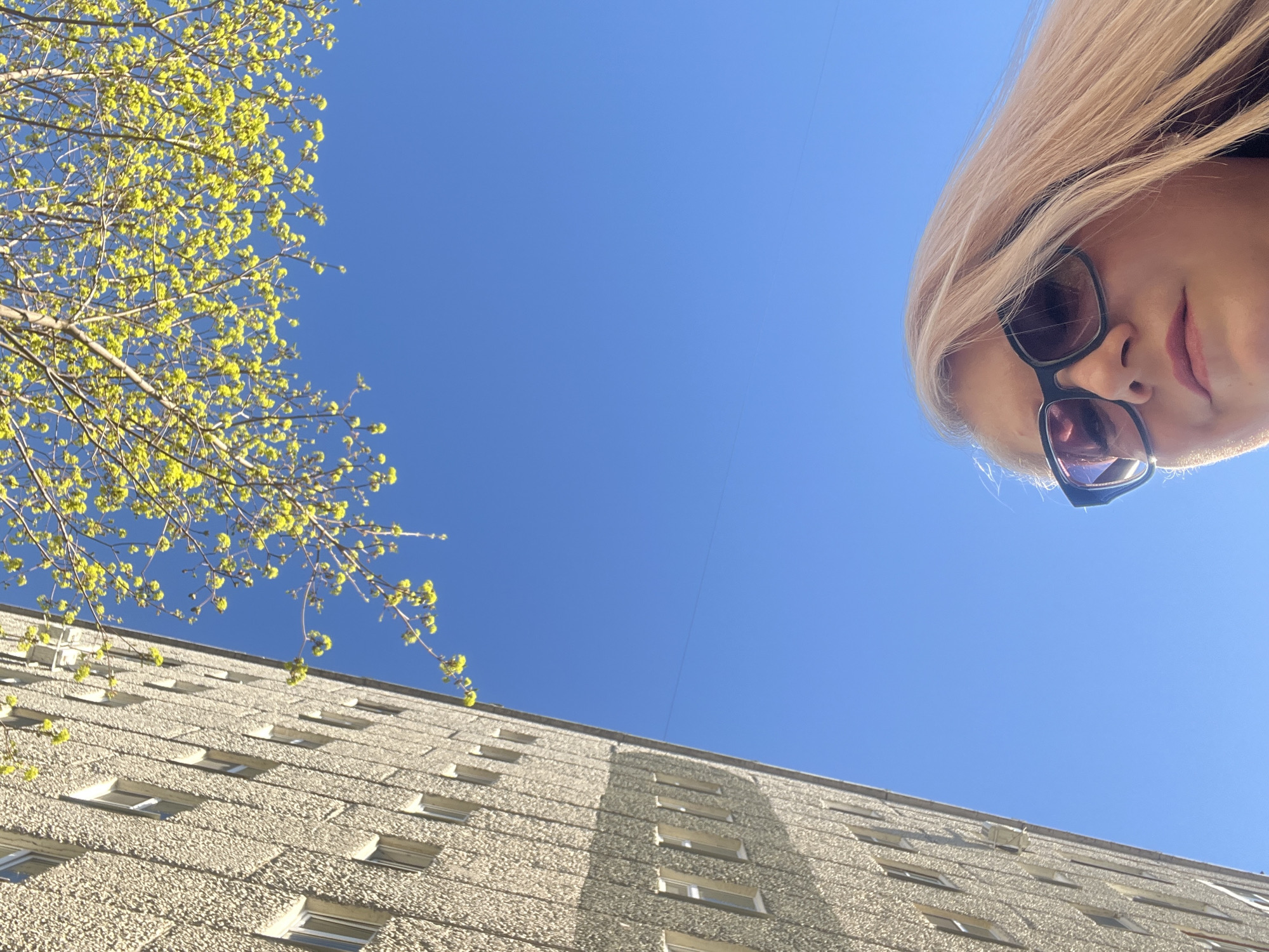 An eye-level, slightly upward shot captures a woman wearing sunglasses from a low angle, positioned to the right side of the frame. Blonde hair obscures part of the clear blue sky behind her. To the left, the frame is filled with tree branches bearing nascent yellow-green leaves. The lower portion of the image features the façade of a multistory building with rows of windows. Based on architectural style, this may be Izhevsk, Russia.