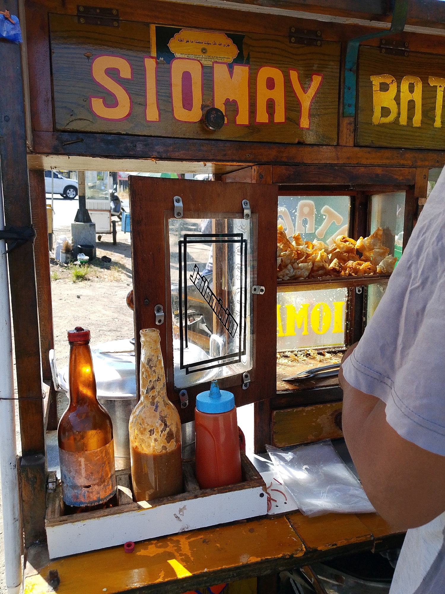 This is a medium shot, eye-level, outdoor photograph of a food cart in Tegalwangi, Indonesia. The cart is made of wood and metal, painted yellow and brown, and appears to be old and well-used. The front of the cart features a display area with two glass panels. The left panel shows a brown glass bottle with a red cap and a partially filled bottle of brown sauce. The right panel displays a small pile of fried food items. Above the display area are two wooden drawers with painted lettering. The left drawer is painted with the word "SIOMAY" in large, orange letters with a pink outline. The right drawer is painted with the word "BAT" in similar orange letters.
Behind the cart, a person's arm and shoulder are visible, wearing a light-colored t-shirt. They appear to be reaching towards the cart. The background shows a dusty street with a white car in the distance and some sparse vegetation. The lighting suggests it is daytime, possibly late morning or early afternoon, with bright sunlight creating shadows. The overall impression is of a casual street food vendor operating in a somewhat dusty, rural or semi-urban environment in Indonesia.