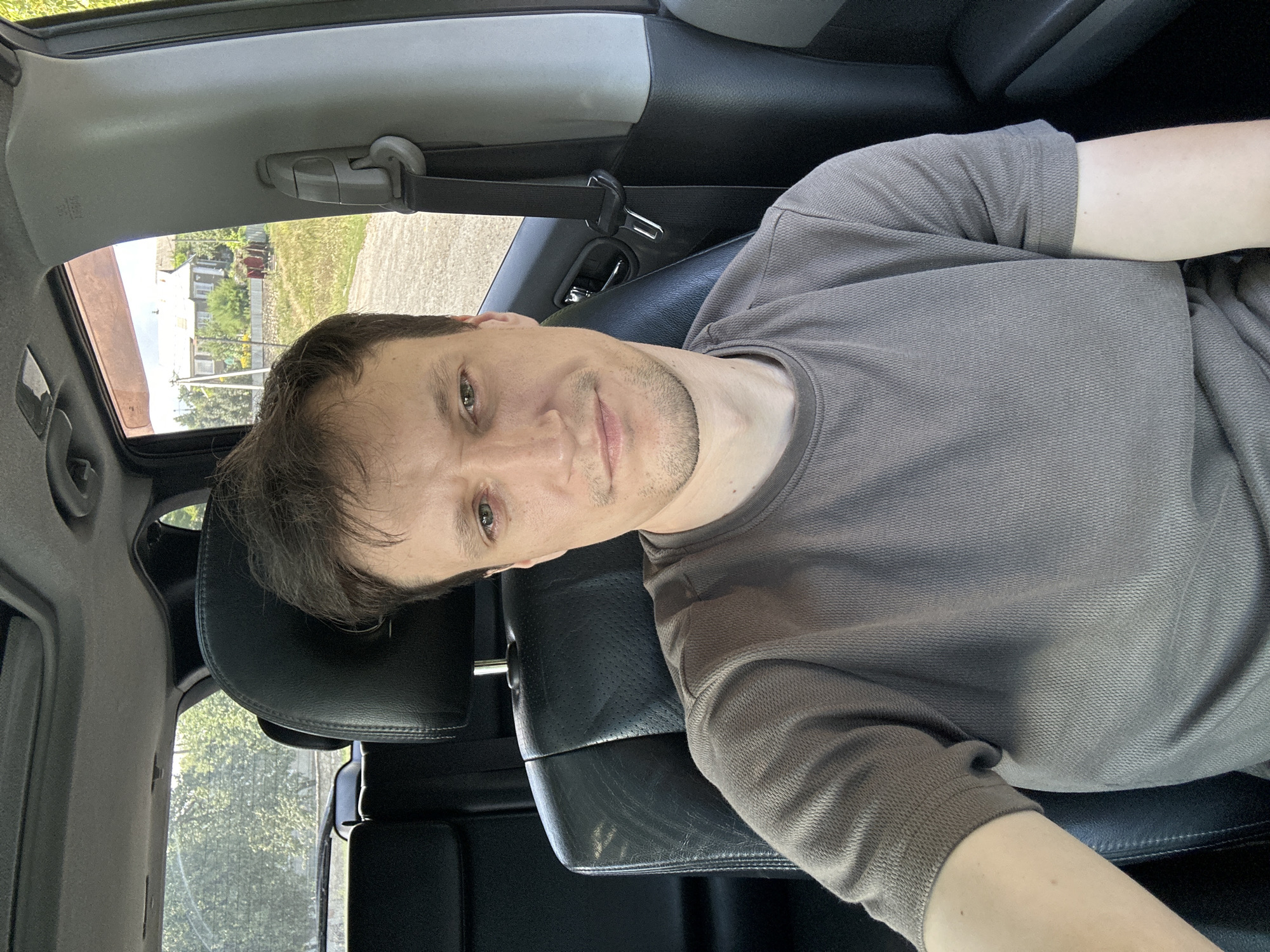 A man is sitting in the driver's seat of a car, taking a selfie. He has short brown hair and is wearing a grey t-shirt. He is looking at the camera with a neutral expression. The car's interior is visible, including the black leather seats and a sunroof. Outside the car, a rural landscape with houses and trees can be seen. The image is taken in Makiv, Ukraine.