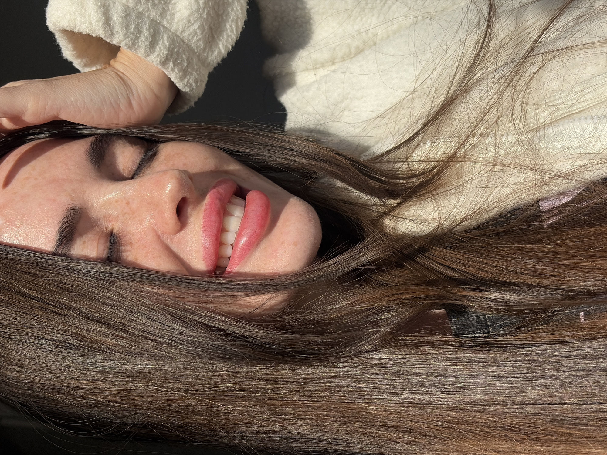 The image displays a close-up portrait of a female subject, approximately mid-20s to early 30s, captured from the chest up. She has long, straight, medium-brown hair. Her eyes are closed, and her mouth is open in a wide smile, revealing both upper and lower teeth. Her lips are full and appear to be colored with a pink-toned product. Freckles are visible across her nose, cheeks, and forehead. Her left hand is raised, with fingers splayed, resting on top of her head, partially obscuring the hair and forehead. She is wearing a cream-colored, textured, fleece-like jacket or sweater, with a dark garment visible underneath near the neck. Strong, direct sunlight illuminates the right side of her face (viewer's left), her extended left arm, and portions of her hair, creating distinct highlights and shadows. The background is a uniform dark grey or black, lacking any discernible features. The specific location within the city of Saint Petersburg, Russia, cannot be determined from the visual information present in the image, as it does not contain any identifiable landmarks or architectural details.