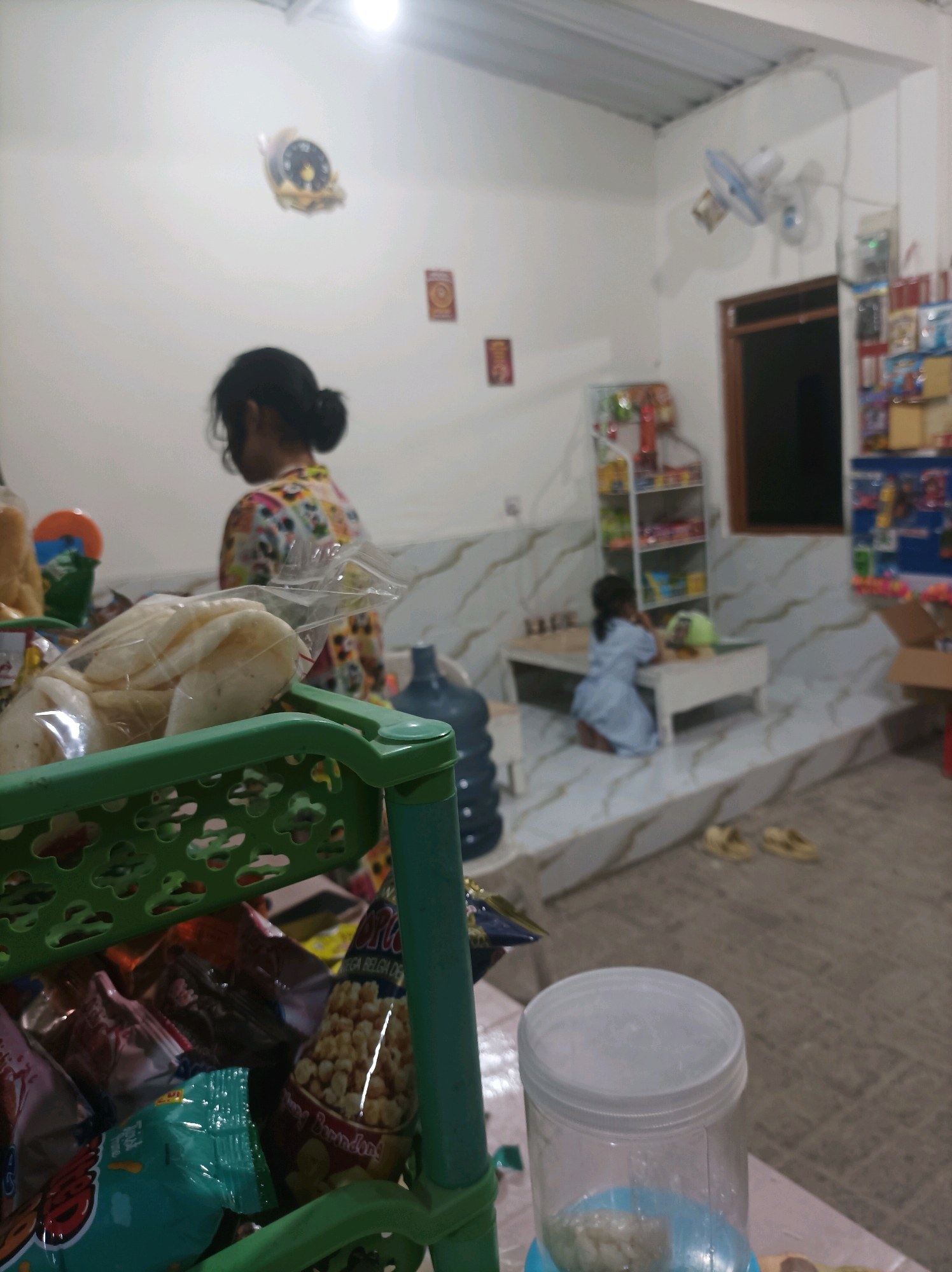 The image depicts an indoor scene, likely a domestic shop or a convenience store, in Canga'an, Indonesia. A young girl wearing a light blue dress is kneeling on a low platform in front of a table, facing a shelf of merchandise. Another person, a woman with dark hair tied back, is standing and facing away from the camera, near a green plastic basket filled with packaged snacks. The environment appears to be a room with white walls, a tiled floor, and a window with dark curtains. There is a wall clock and a fan mounted on the wall. A large blue water jug is visible in the background, along with shelves stocked with various products, including snacks and what appears to be household items. A partially visible jar with a white lid is in the foreground. The lighting suggests it is either evening or an indoor setting with artificial light.