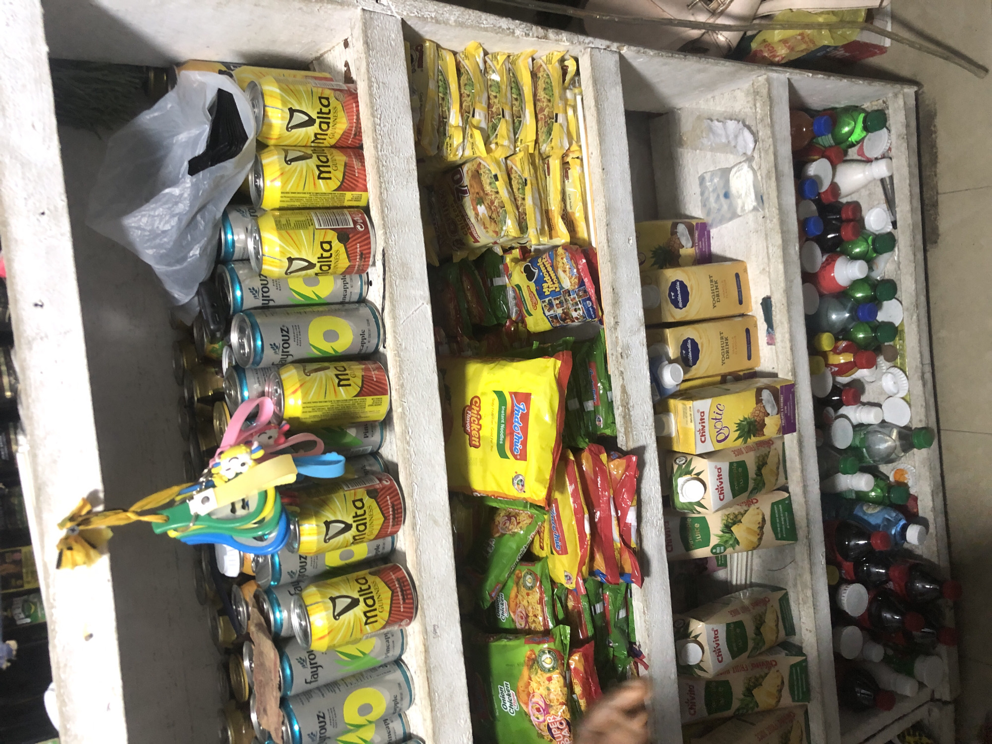 The image shows a cluttered shelf display in what appears to be a small shop or market stall in Ado, Nigeria. The shelves are stocked with a variety of food and beverage items.
On the top shelf, there are numerous cans of drinks, including what appears to be Malta Guinness and Loya. Some cans are light grey with green accents, and others are yellow and red. A bunch of colorful keychains hangs from the edge of the shelf. Behind the cans, a white plastic bag is bunched up.
The middle shelf is filled with packets of instant noodles and small juice cartons. There are yellow packets of Indomie noodles, some with "Chicken Flavor" and "Super Pack" visible. There are also smaller, individual packets of noodles. Towards the right, there are green and yellow cartons of "Chivita Pineapple Juice" and "Chivita Exotic".
The bottom shelf contains an assortment of plastic bottles of drinks. These bottles have various colored caps and liquids, including dark red, blue, and green. Some bottles appear to be sodas or other carbonated beverages. There are also a few loose bottle caps scattered on the shelf.
The shelves themselves appear to be made of wood and are somewhat aged, with some discoloration and wear. The overall environment suggests a retail setting where items are displayed for sale.