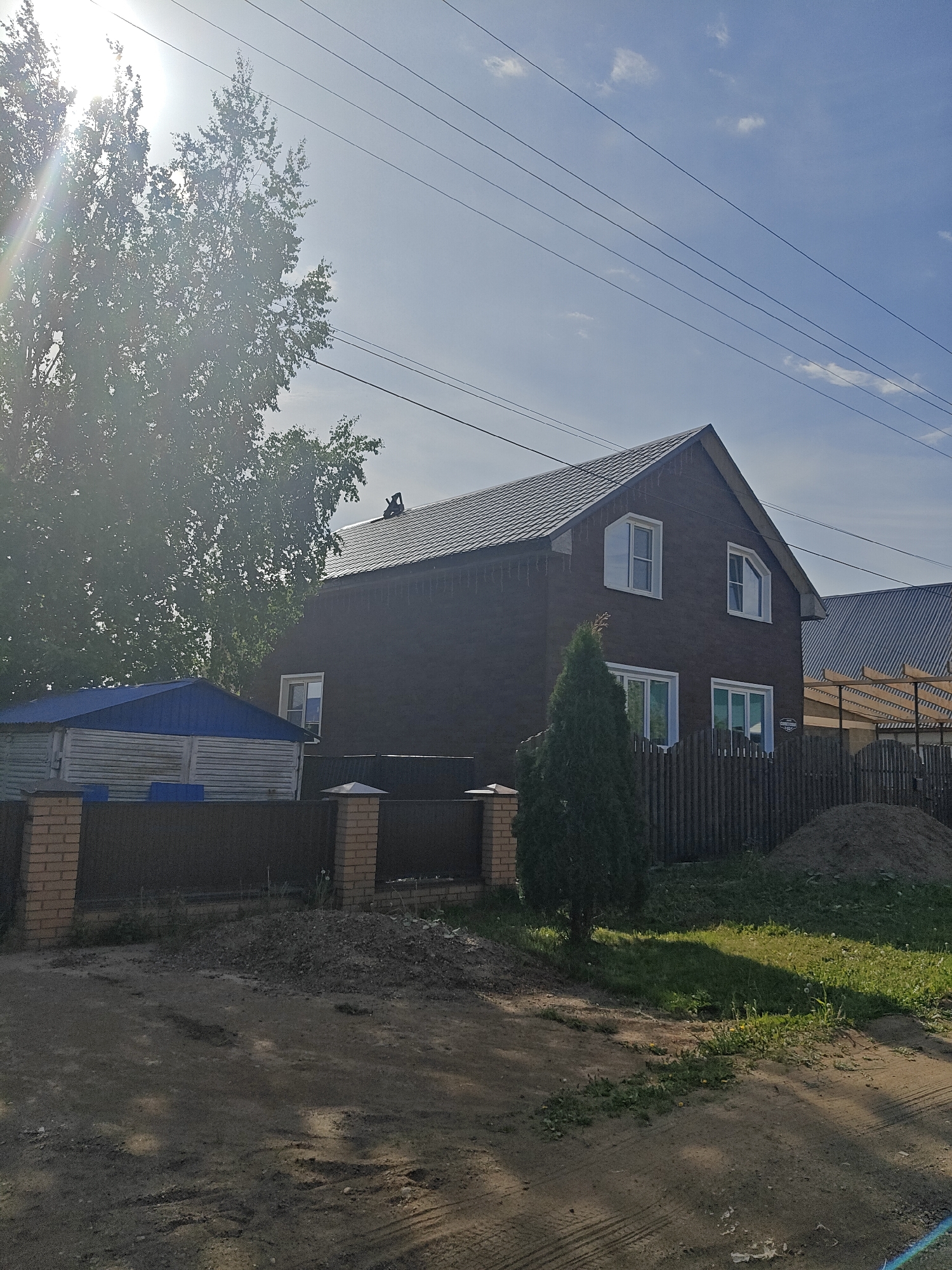 A photograph taken in Фоки, Russia, shows a two-story house with dark brown siding and a gray tiled roof. Two windows are visible on the upper floor and two on the ground floor, all framed in white. A small evergreen tree stands in the yard near a wooden fence. To the left of the house, there's a blue-roofed shed behind a brick and metal fence. A pile of dirt is visible near the fence to the right of the house. Power lines stretch across the sky. The sun is shining brightly, creating a glare, and casting shadows across a dirt road in the foreground. A person appears to be on the roof of the house. A large tree with dense foliage is on the left side of the frame.
