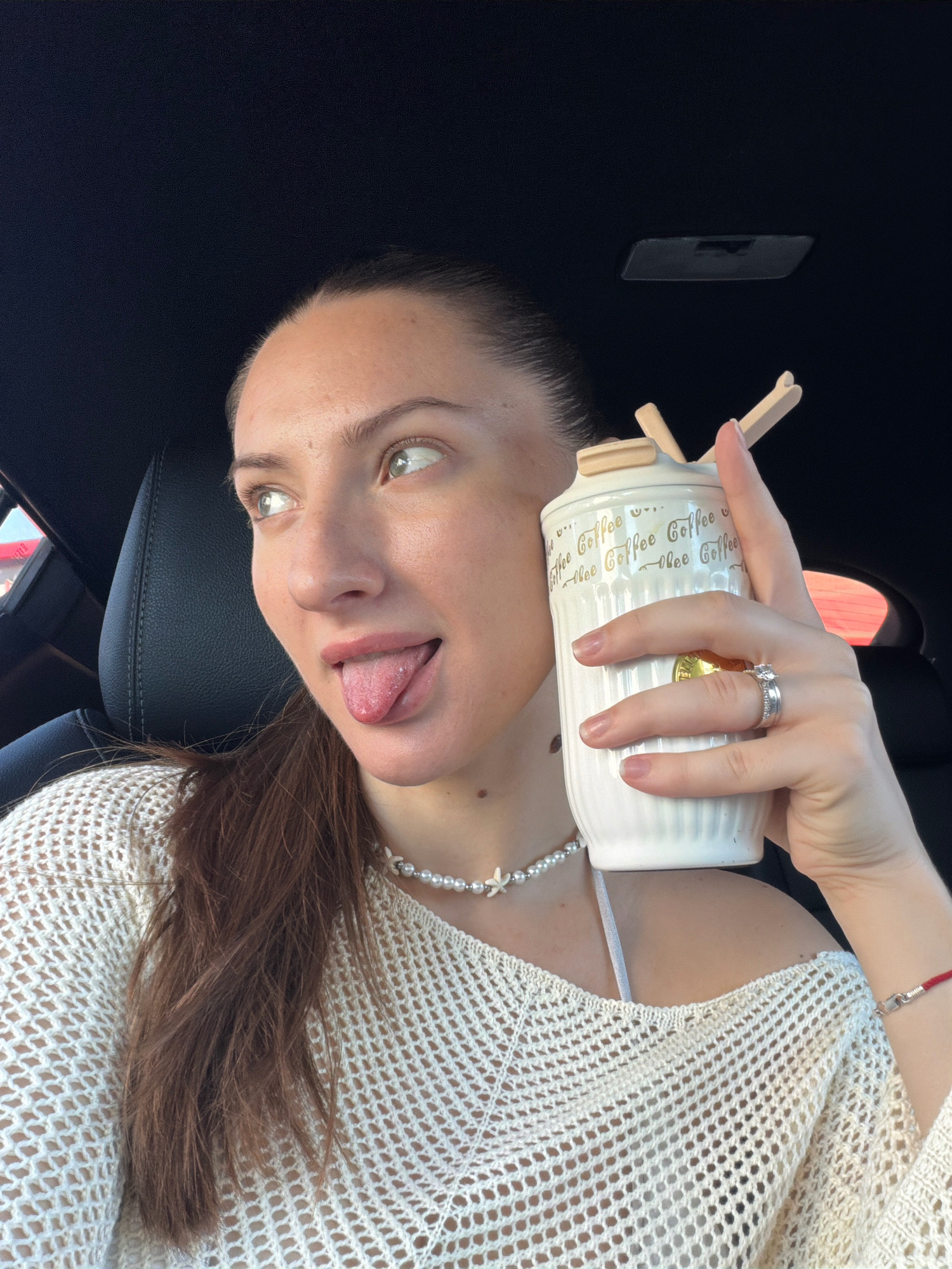 A young woman with brown hair pulled back is in the driver's seat of a car, possibly in Sharm El Sheikh, Egypt. She is wearing a white crocheted top and a pearl necklace. Her tongue is sticking out and her eyes are looking to the side. She is holding a white ceramic coffee cup with a lid and two wooden sticks. The car's interior is visible, including a black leather seat and the roof. A red string bracelet is visible on her right wrist.