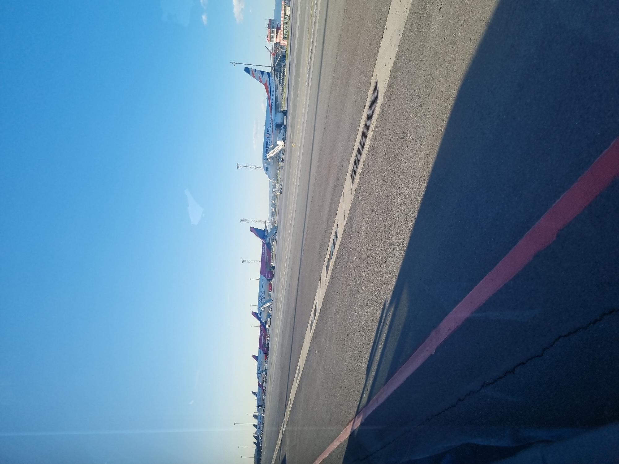 The image captures a view from an elevated position, possibly inside a vehicle, overlooking an airport tarmac. Several commercial airplanes are parked in a row, with at least one having a boarding staircase attached. The sky is clear and blue. The tarmac is gray and marked with white and red lines. A shadow obscures a portion of the foreground. Based on the presence of Smartwings airplanes, the location is likely Sofia Airport in Sofia, Bulgaria. No people are visible in the image.