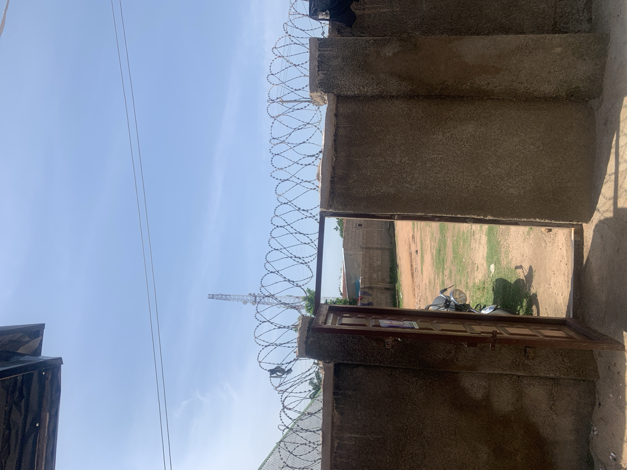 The image shows an open gateway with a barbed wire fence along the top of the wall. A dark-colored motorcycle is parked behind the open gate on the right side of the image. The ground is a mixture of dirt and patches of green grass. In the background, there are several buildings constructed from concrete blocks. A tall telecommunications tower is visible in the distance against a clear blue sky. A portion of a dark-colored roof structure is visible in the upper left corner of the image. This scene is located in Jauro Yinu, Nigeria.