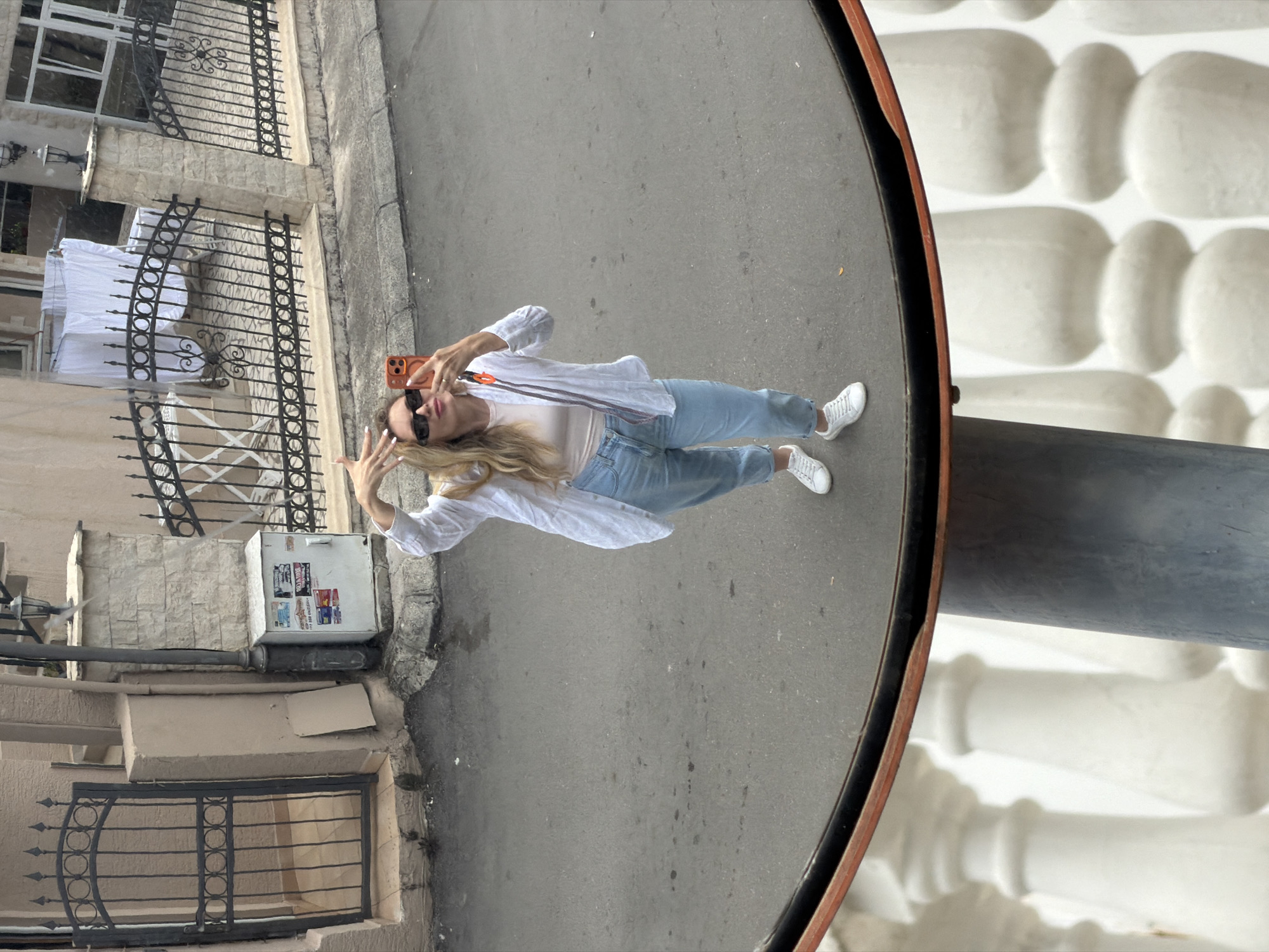 A medium, upward-angle, eye-level shot shows a woman reflected in a convex mirror in Budva, Montenegro. The woman, wearing sunglasses, a white long-sleeved shirt over a light-colored t-shirt, and light blue jeans, holds an orange phone and makes a V-sign with both hands. She has long blonde hair. The mirror reflects a street scene with sidewalks, wrought-iron fences, and a utility box. The background behind the mirror reveals a white textured wall. The street surface appears to be asphalt.