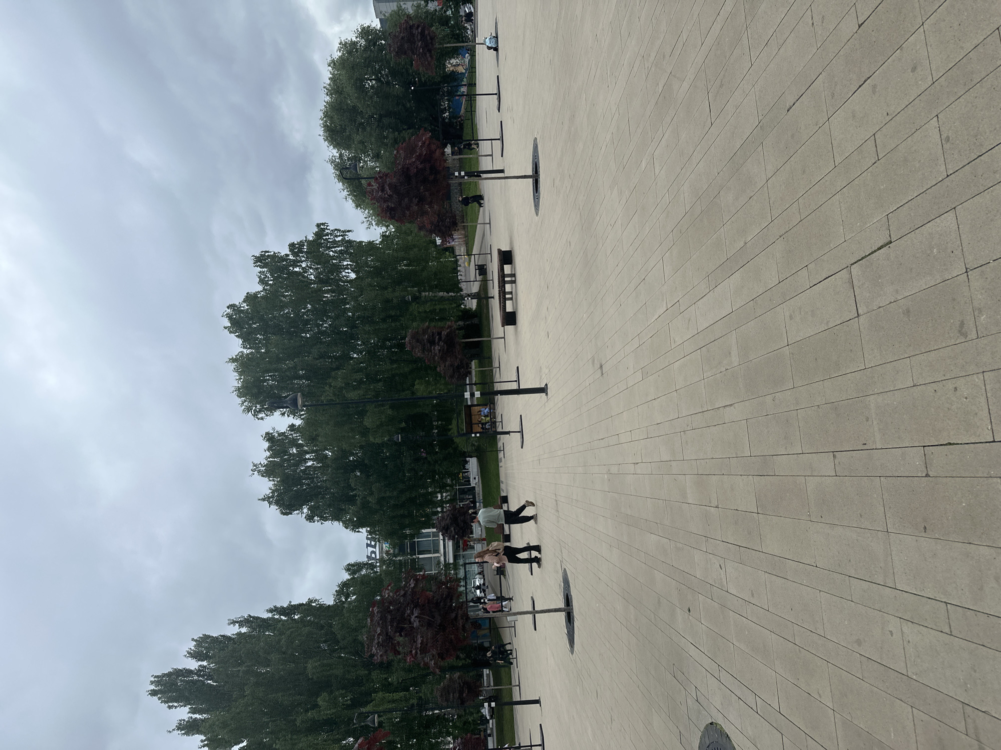 The image depicts a public plaza in Izhevsk, Russia, situated in front of the Kinotsentr "Molodyozhny" cinema, as indicated by the partial Cyrillic lettering "ЪН" visible on the main building in the background. The scene is observed under an overcast sky with light grey clouds, suggesting daytime conditions.
The plaza is paved uniformly with light beige or grey rectangular concrete tiles laid in a staggered pattern. A circular metal manhole cover is partially visible in the lower-left foreground. Approximately nine small, ornamental deciduous trees with dark reddish-brown foliage are symmetrically spaced across the plaza, each planted within a circular metal grate. Numerous tall, black lampposts with single light fixtures are interspersed among the trees. One dark-colored public bench with a backrest is centrally located in the mid-ground.
In the background, dense clusters of tall, mature green deciduous trees flank the main building. The multi-story building, identified as the Kinotsentr "Molodyozhny," features a light-colored facade with large windows. To the right of this building, a low wall or structure displays a colorful mural.
Several individuals are present in the plaza. In the mid-ground, a woman wearing a light-colored top and dark pants, carrying a light backpack, walks away from the viewer alongside a man in a light green long-sleeved top and dark pants. In the left background, multiple indistinct figures are walking, one appearing to push a stroller. On the right side of the background, two or three individuals are seated on the ground or a bench near the mural. Individuals are engaged in walking and sitting, with no specific interactions clearly discernible.