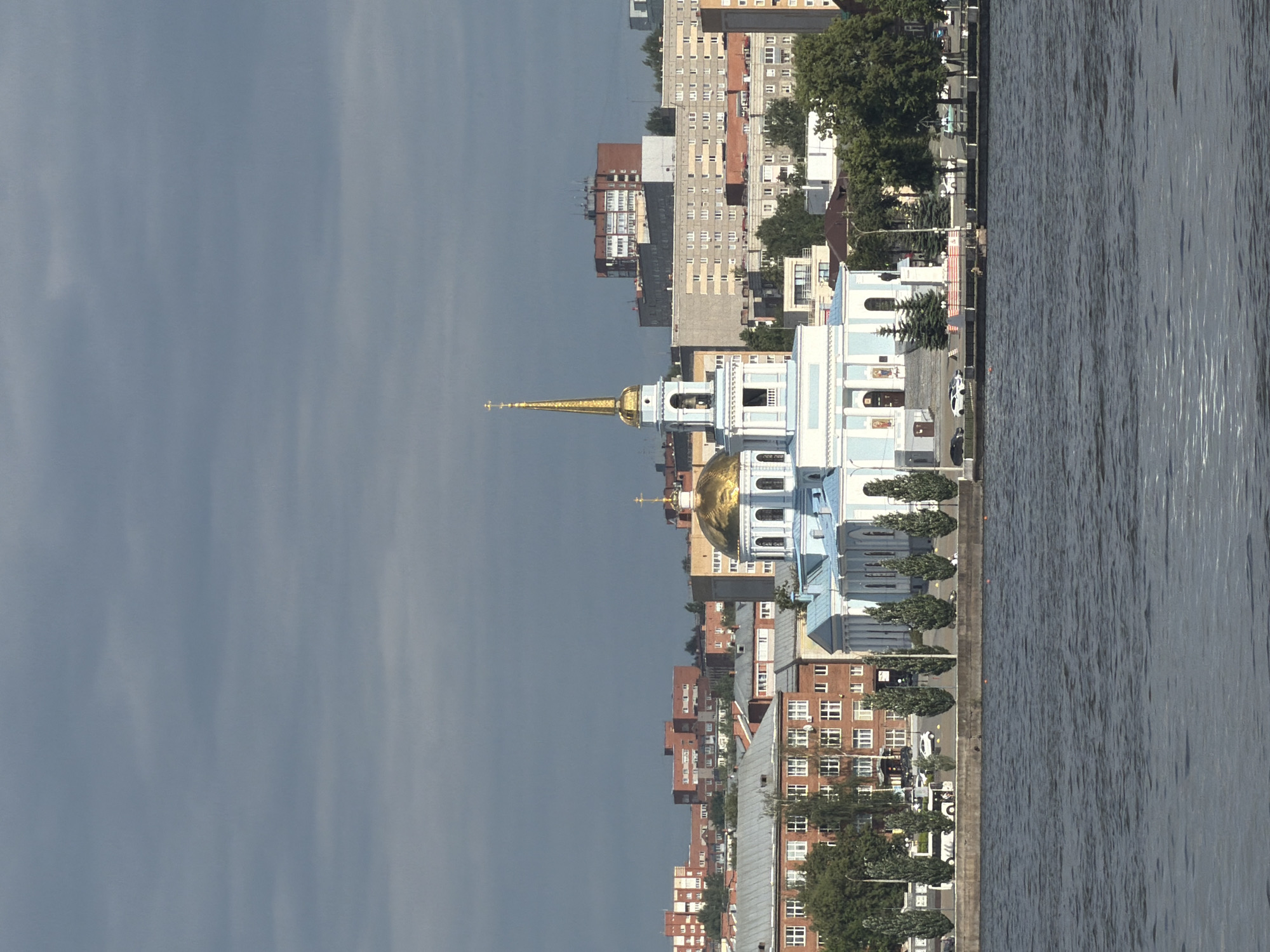 This image depicts a panoramic view of the city of Votkinsk, Russia, focusing on the Church of the Transfiguration (Preobrazhenskaya Tserkov) and its surrounding urban landscape, as seen from across the Votkinsk Pond.
In the middle ground, slightly right of center, stands a large, light-blue and white Orthodox church. It features a prominent golden onion dome on its main structure and a taller, slender golden spire topped with an Orthodox cross on its bell tower, both reflecting sunlight. The church exhibits classical architectural elements, including columns, arched windows, and multiple tiers. Steps lead up to its main entrance.
In the foreground, a wide, dark blue-grey body of water, consistent with the Votkinsk Pond, occupies the lower third of the image. The water surface shows subtle ripples. A concrete embankment separates the water from the land. Along this embankment, a paved area or road is visible, on which a white bus and several white cars are parked or moving. A row of tall, slender evergreen trees lines the embankment, interspersed with other green foliage. Small human figures are discernible near the embankment and on the steps of the church, though their specific details are indistinct due to the distance.
The background consists of a dense urban environment with numerous multi-story buildings. To the left of the church, older reddish-brown brick buildings with numerous rectangular windows are visible. To the right and further back, a mix of residential and commercial structures, varying in color (grey, beige, brick red), form the city skyline. Some buildings feature flat roofs with visible utility structures.
The sky, occupying the upper half of the image, is overcast with layers of grey-blue clouds, indicating diffused lighting conditions.