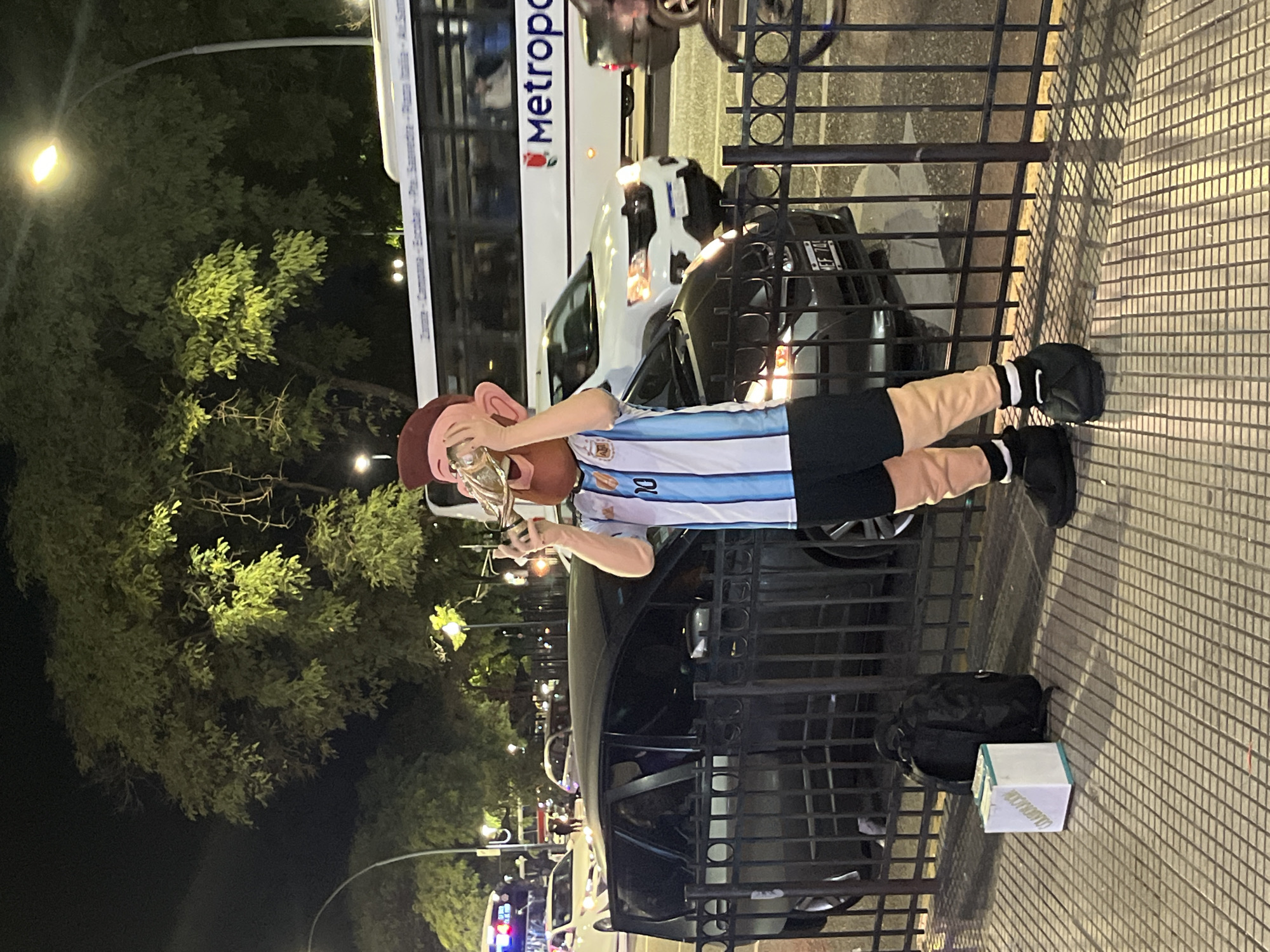 A person in an Argentina soccer jersey and a mask of Lionel Messi stands on a city sidewalk at night, next to a metal fence. The person holds a replica of the FIFA World Cup trophy. A bus is parked behind them with "Metropol" written on the side. There are cars on the road next to the bus. Streetlights illuminate trees in the background. The location is likely in Buenos Aires, Argentina, based on the jersey and mask indicating association with the country. A backpack and a white box rest on the sidewalk. The person in the Messi mask appears to be posing or performing for onlookers.