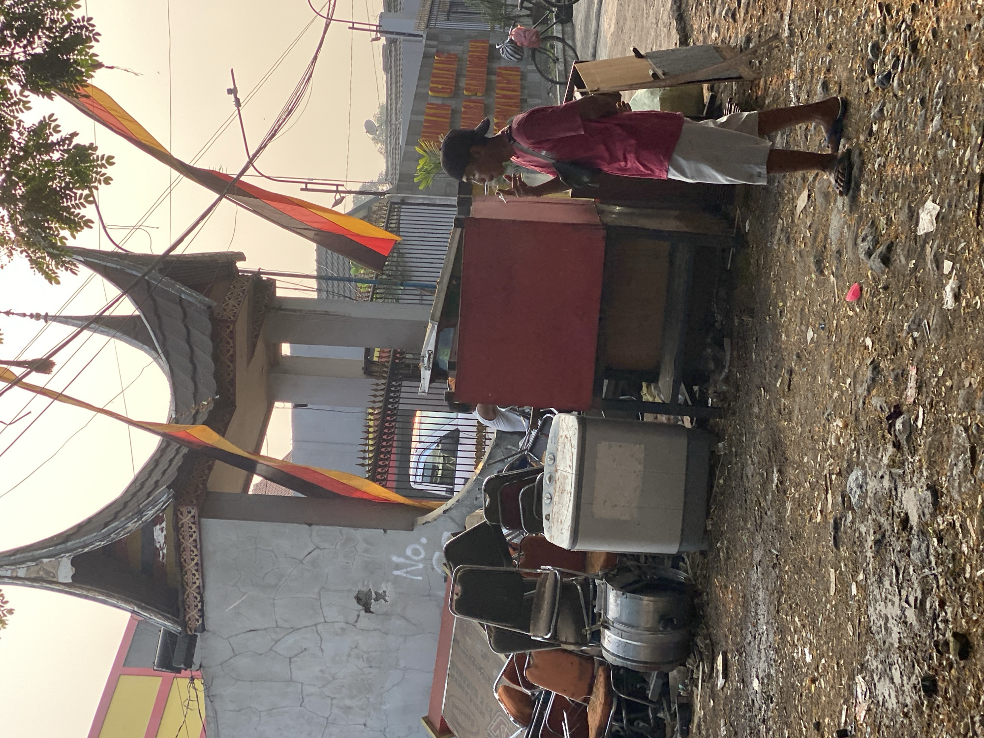 A young man stands on a dirty lot in Kasihan, Indonesia, smoking a cigarette. He wears a pink t-shirt, white shorts, and flip-flops. Behind him, a pile of old chairs and a washing machine are strewn on the ground. A red wooden stall is to his left, and in the background, a building with traditional Indonesian architecture is visible, along with flags and power lines. The overall scene suggests a somewhat neglected urban environment.