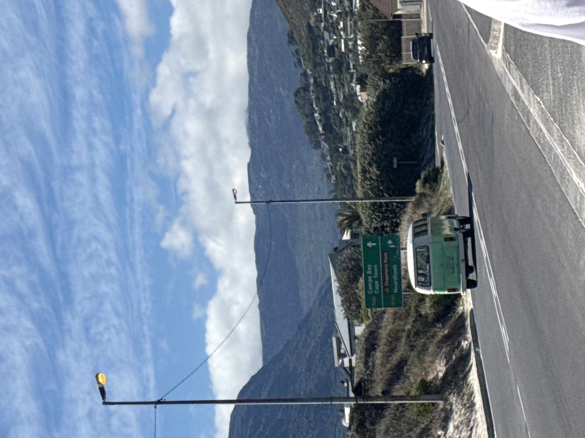 A sunlit view in Hout Bay, South Africa, showcases a road running along the coast with a mountainside in the background partially obscured by clouds. A green and white vehicle is positioned on the road, adjacent to a signpost directing towards Camps Bay, Cape Town, Chapman's Peak, and Noordhoek. A light pole stands on the left side of the image, and there are buildings visible further along the mountain.