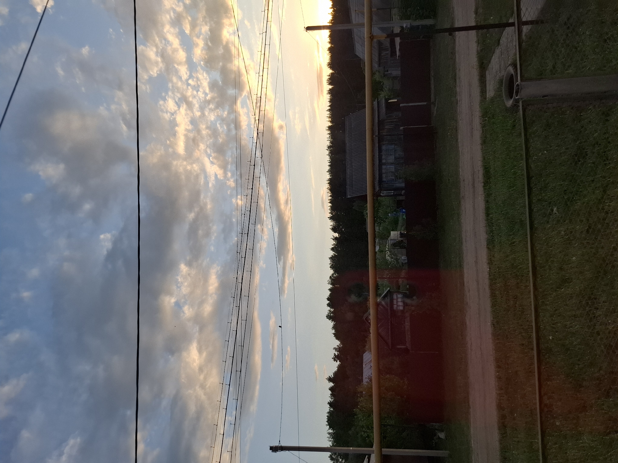 The image depicts a rural scene in Амзя, Russia, at what appears to be dusk or dawn. The sky is filled with clouds illuminated by a low sun, with power lines extending across the view. A dirt road runs along the right side of the image, bordered by vegetation and various structures, including houses and outbuildings. The scene evokes a quiet, countryside atmosphere.