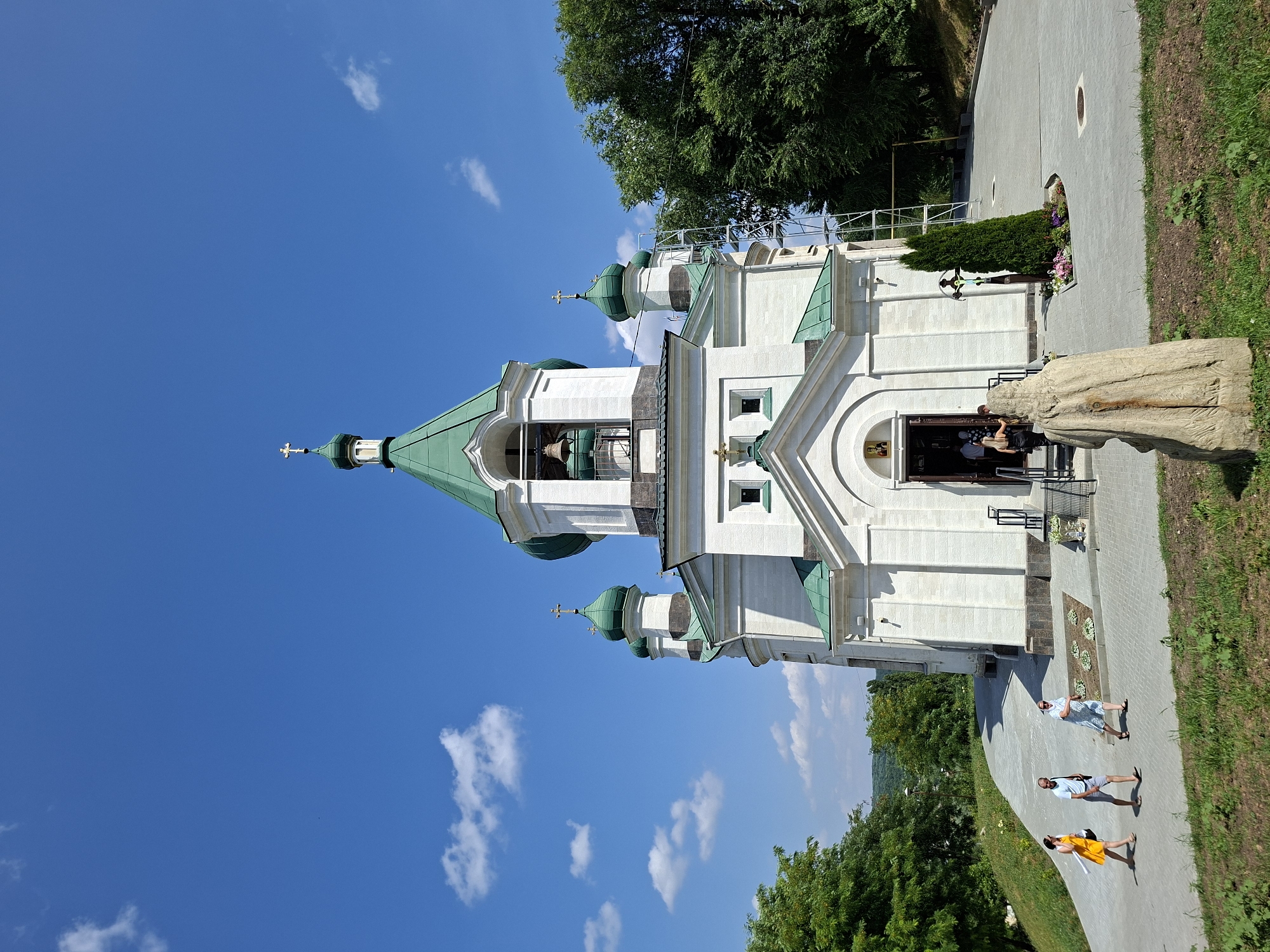 The image depicts the exterior of a white church with green domes, located in the city of Condrița, Moldova. The church features a central bell tower with a large bell and a cross at its apex. Several smaller domes with crosses are also visible. The church entrance is open, and a few people are seen entering and exiting.
In the foreground, a rough-hewn wooden sculpture stands. To the left of the church, a paved pathway leads up to the entrance, with a grassy slope beside it. Three individuals are walking on the path, dressed in casual attire. To the right of the church, a tall evergreen tree stands next to a small flower bed with a cross. Beyond the church, lush green trees form the background, with a hilly landscape visible in the distance. The sky is a clear blue with a few scattered white clouds. Scaffolding is erected near the side of the church.