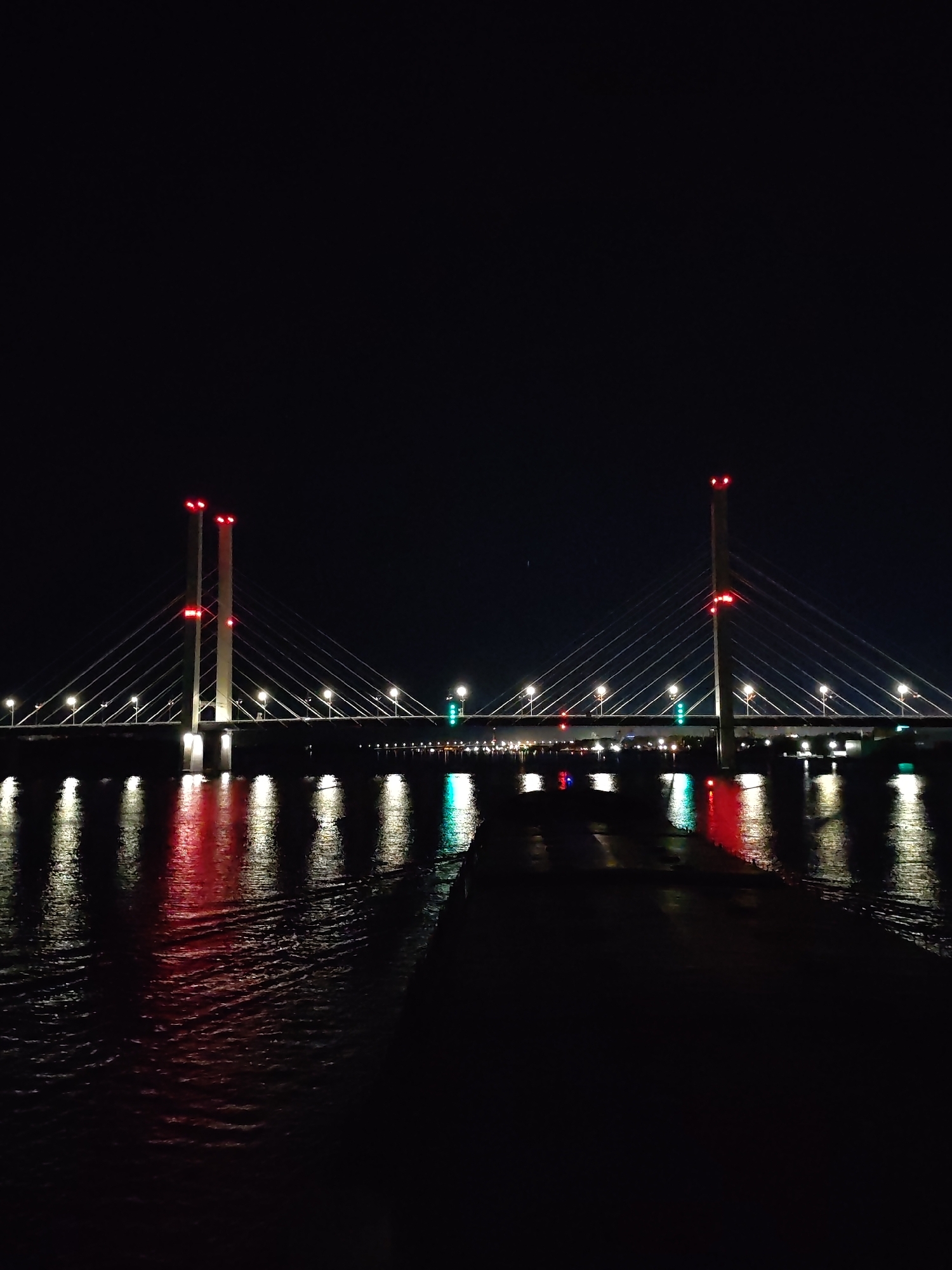 The image shows a cable-stayed bridge at night. The bridge has two towers with red lights on top. There are several street lights on the bridge. The lights from the bridge are reflected in the water below. The city is Unknown, Unknown. A dark pier stretches into the lower portion of the water.