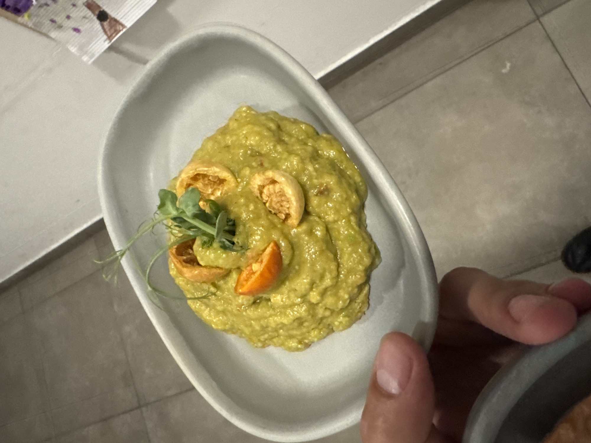 A close-up shot captures a light grey, oval-shaped dish filled with a vibrant green, mashed substance, possibly guacamole or a similar dip. The dish is garnished with three small, round orange pieces, likely dried fruit or halved cherry tomatoes, and a sprig of delicate green microgreens. A hand is visible at the bottom left, holding the dish. The background appears to be a tiled floor with a slightly blurred white surface to the right, suggesting an indoor setting, possibly a restaurant. No specific city or location is discernable from the visual cues.