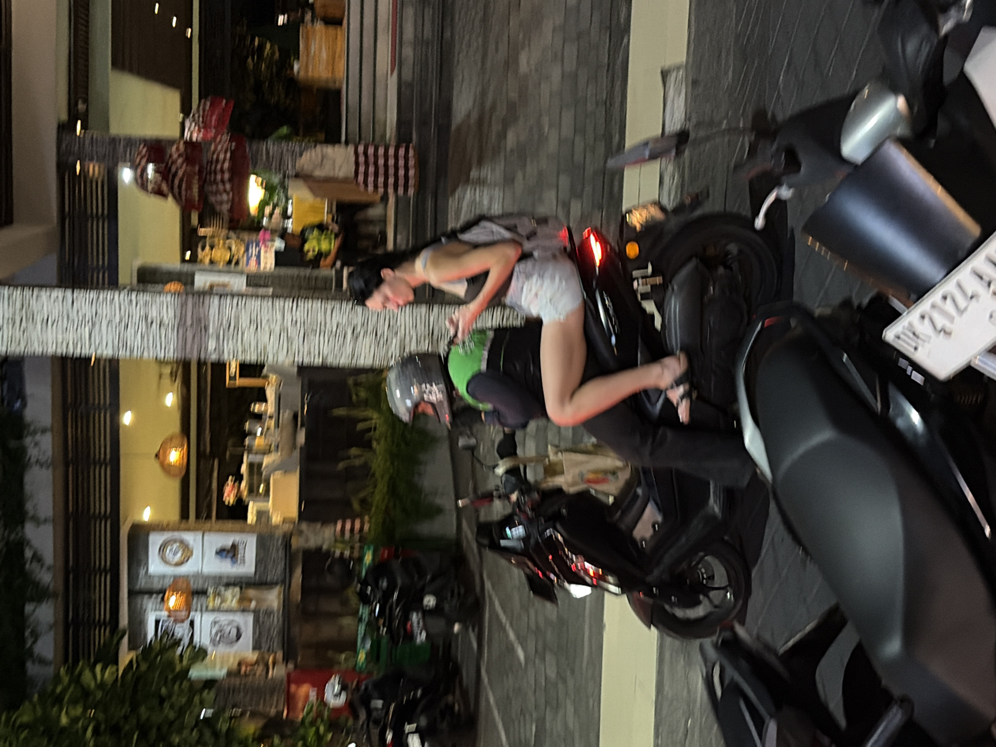 In Kuta, Indonesia, at night, two people are riding a black scooter. The person in the front, wearing a green vest and a helmet, appears to be driving. The person in the back, a woman with long dark hair, is wearing a gray tank top and white shorts, and is looking at her phone. She has a gray backpack on. The scooter has a canvas bag hanging from the handlebars. In the background, there is a building with lights and some decorations, possibly a restaurant or shop. Other scooters are parked nearby. The ground is paved with interlocking blocks.