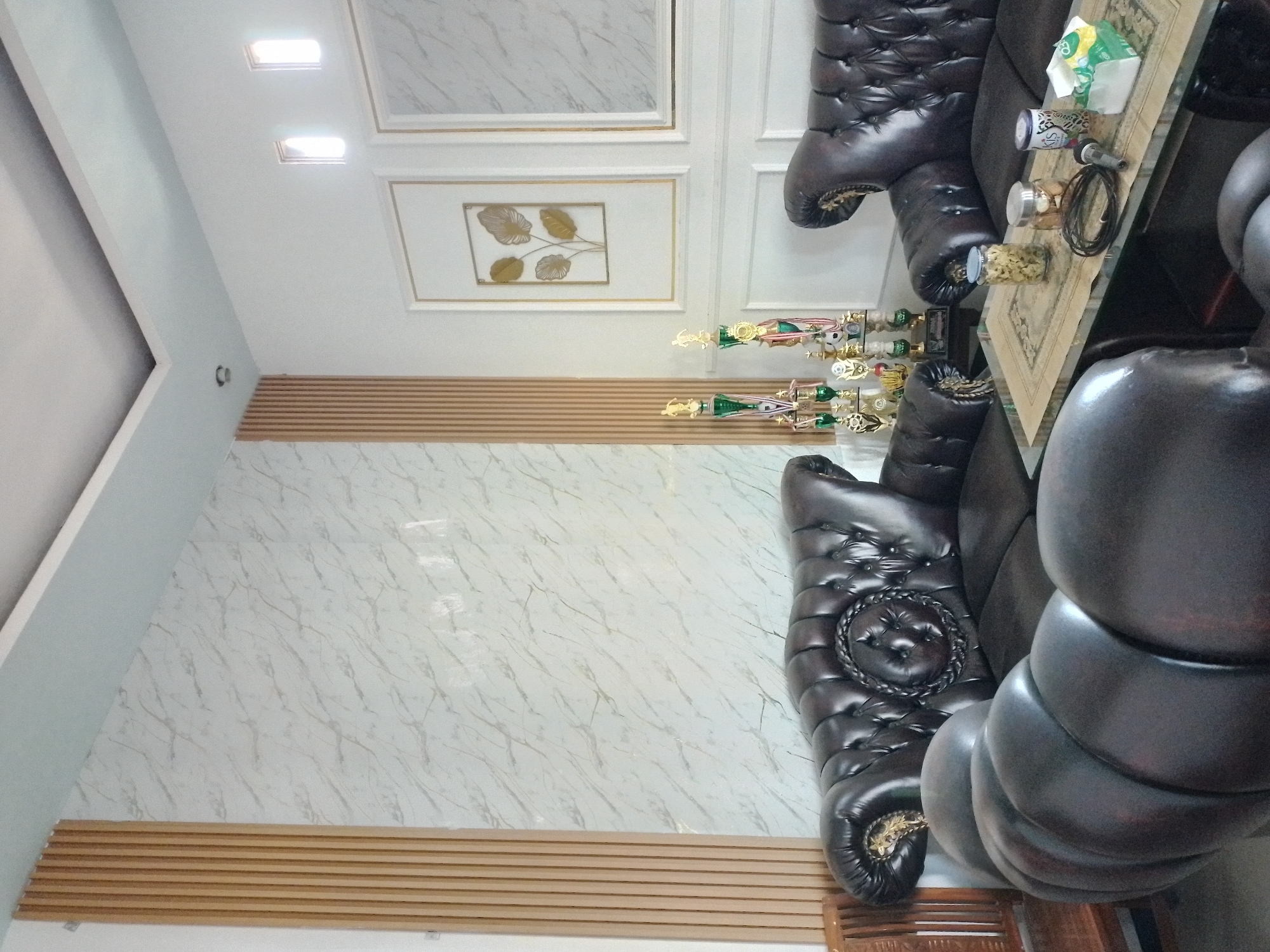 The image depicts an indoor scene, likely a living room or reception area, in Ngancar, Indonesia. Two dark brown leather Chesterfield sofas are the prominent furniture pieces. The sofa on the left is larger and L-shaped, with tufted upholstery and decorative trim. The sofa on the right is a smaller, two-seater version. In the foreground, a glass-topped coffee table displays several objects: two glass jars containing snacks, a coiled black cable, a microphone, and a box of tissues. Behind the coffee table, a collection of trophies of varying sizes and designs is arranged on a shelf. The walls are decorated with a marble-patterned wallpaper and vertical wooden slatted panels. Two framed pieces of art, featuring botanical motifs, are mounted on the wall. The ceiling is white and features recessed lighting. Two small, illuminated windows are visible high on the wall. The overall impression is one of a well-furnished and maintained interior space. There are no people visible in the image.