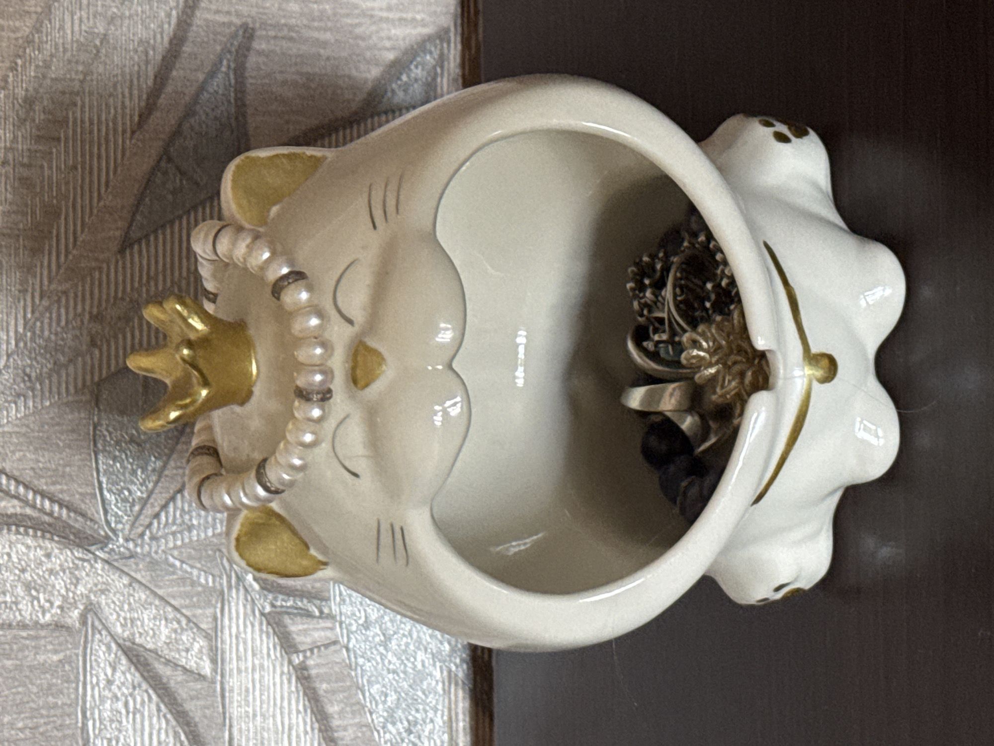 An overhead shot shows a white ceramic cat-shaped trinket dish on a dark brown surface, likely a table or shelf. The dish is adorned with a gold crown on its head, a pearl necklace around its neck, and gold accents on its ears and paws. Inside the dish, a collection of jewelry, including rings and necklaces, is visible. The background features a textured wallpaper with a grey, abstract pattern.