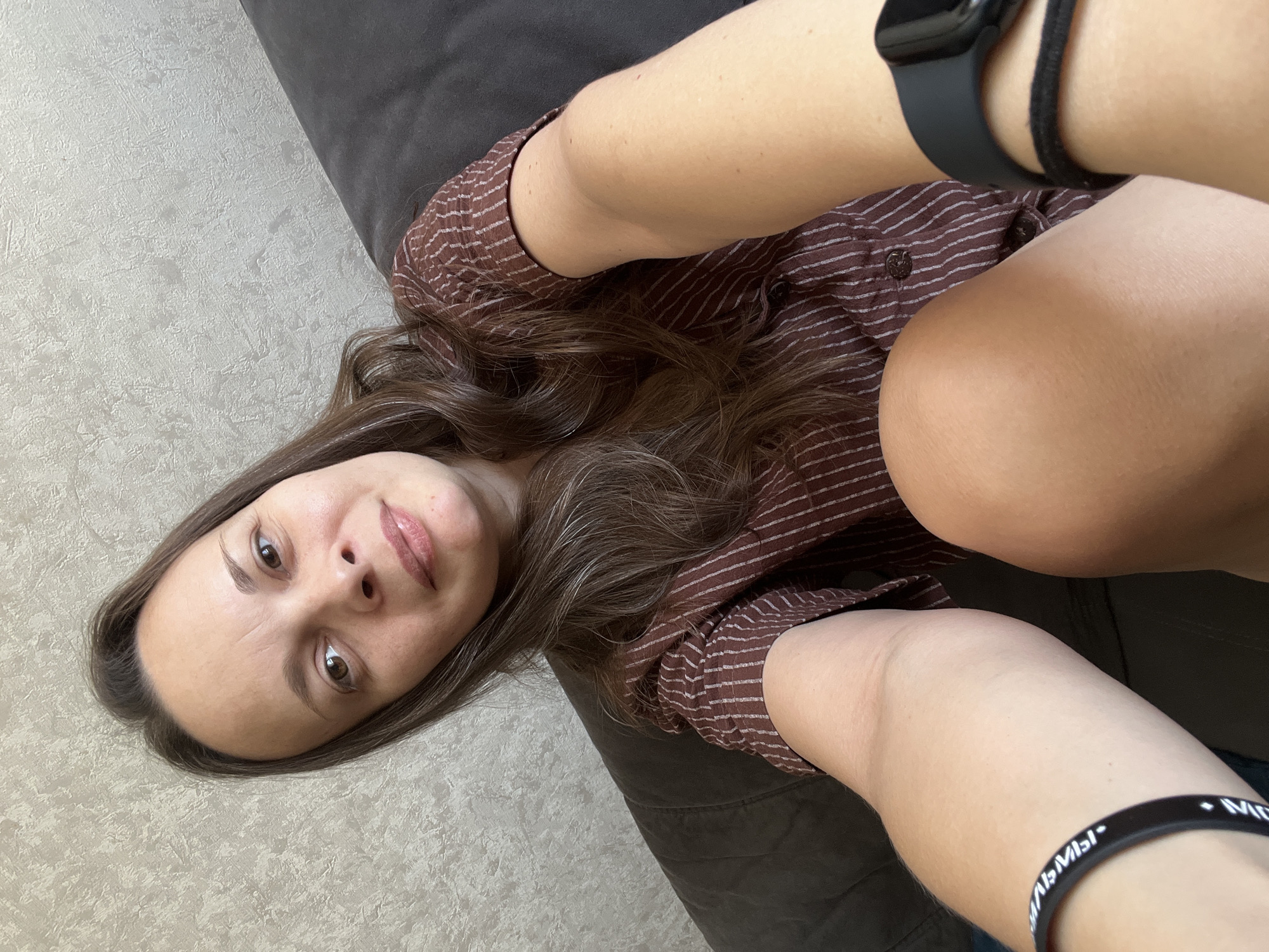The image shows a woman sitting on a dark couch in what appears to be a living room. She is looking at the camera and smiling slightly. She has long, brown hair and is wearing a brown, striped shirt. On her left wrist, she is wearing a black bracelet with white text. On her right wrist, she is wearing a black smartwatch with a black band. The background is a textured, light-colored wall. The scene is illuminated by natural light. There is no information about the location within the city of Tolyatti, Russia.