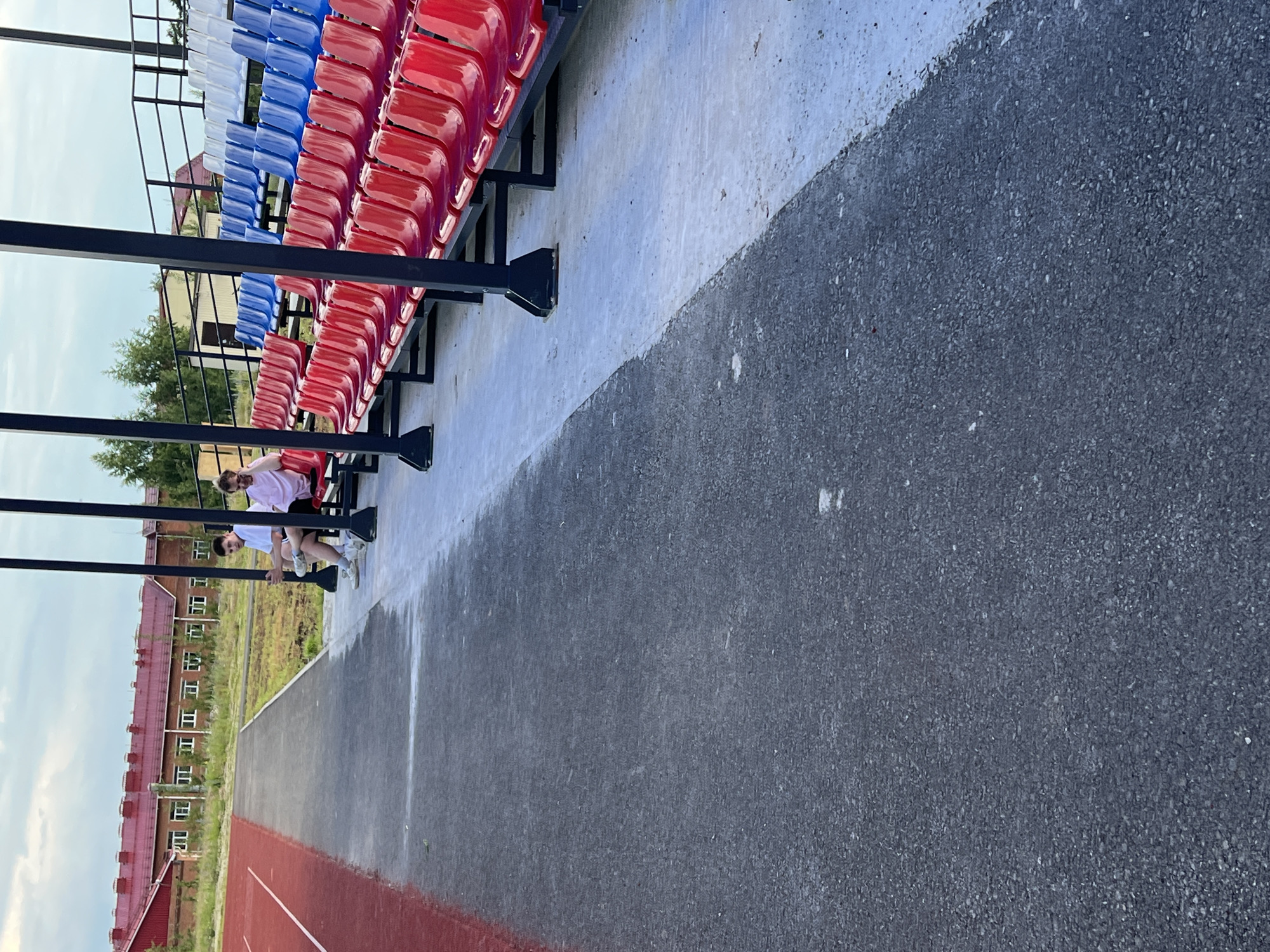 The image shows an outdoor stadium with bleachers. The bleachers have red, white, and blue seats, arranged in rows. Two people are visible sitting on the bleachers, near the bottom of the frame. One person, wearing a pink shirt and black shorts, is leaning back. The other person, wearing a white shirt and black shorts, is sitting beside them. In the background, there is a building with a red roof and several windows. There is also a running track, with a red outer edge. The sky is overcast, suggesting it might be a cloudy day. The image appears to have been taken during the daytime. The scene is set in Noviy, Russia.