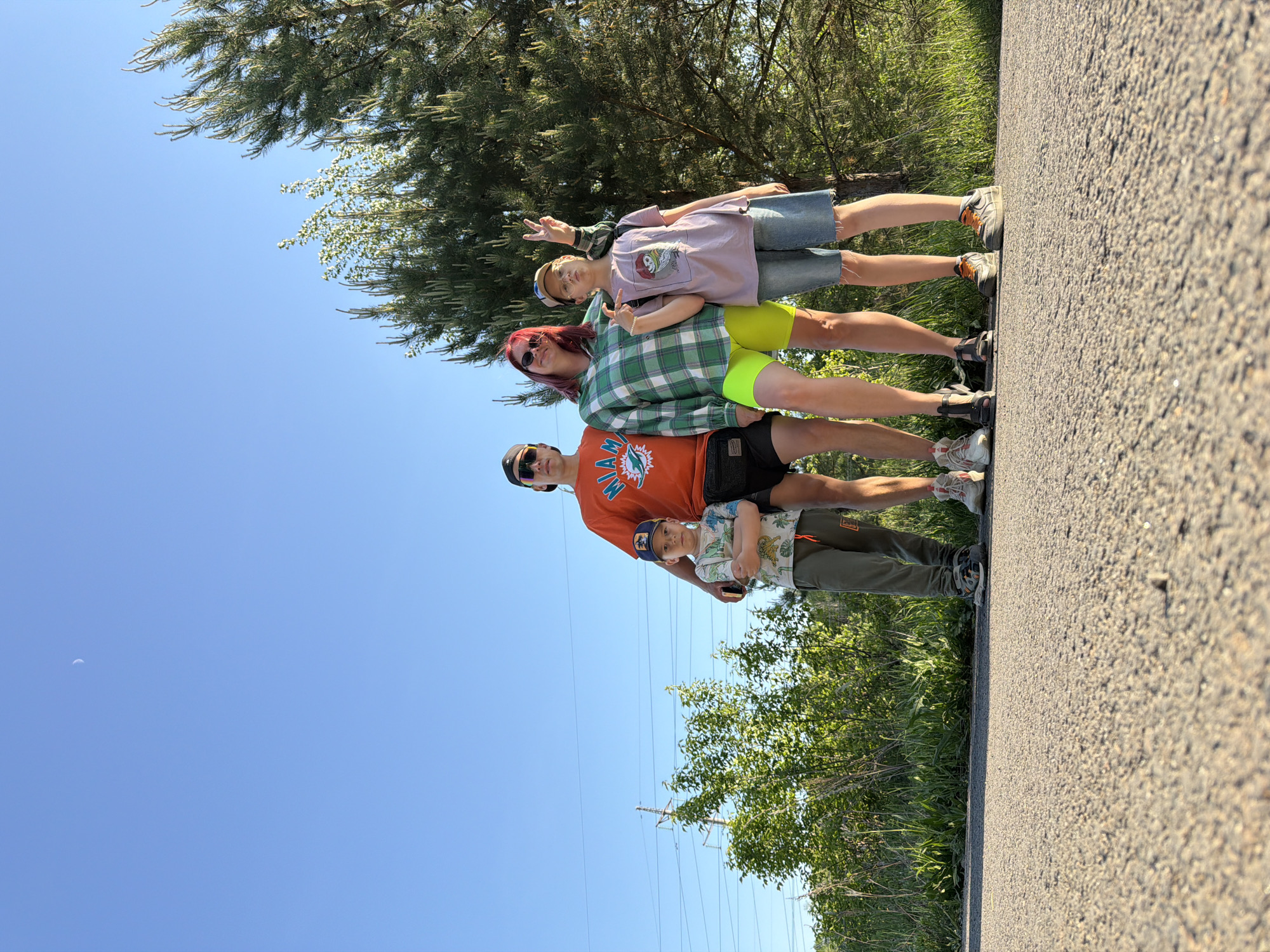 Four individuals stand in a line on a paved surface, likely a road or pathway, in an outdoor setting. The first person in line is a young boy wearing a baseball cap, a short-sleeved shirt with a pattern, and olive green pants. The second is a man wearing sunglasses and an orange T-shirt with a sports team logo. Next is a woman with red hair and sunglasses, wearing a green and white plaid shirt. The final person is a teenager with short hair, wearing a pink shirt and denim shorts and flashing a peace sign. Trees and vegetation are visible in the background, with a clear blue sky overhead and a faint crescent moon in the upper left corner. High-voltage power lines and towers are noticeable at the horizon. This image was likely taken in a public space, possibly near Chaikovsky, Russia. The photo has been rotated.