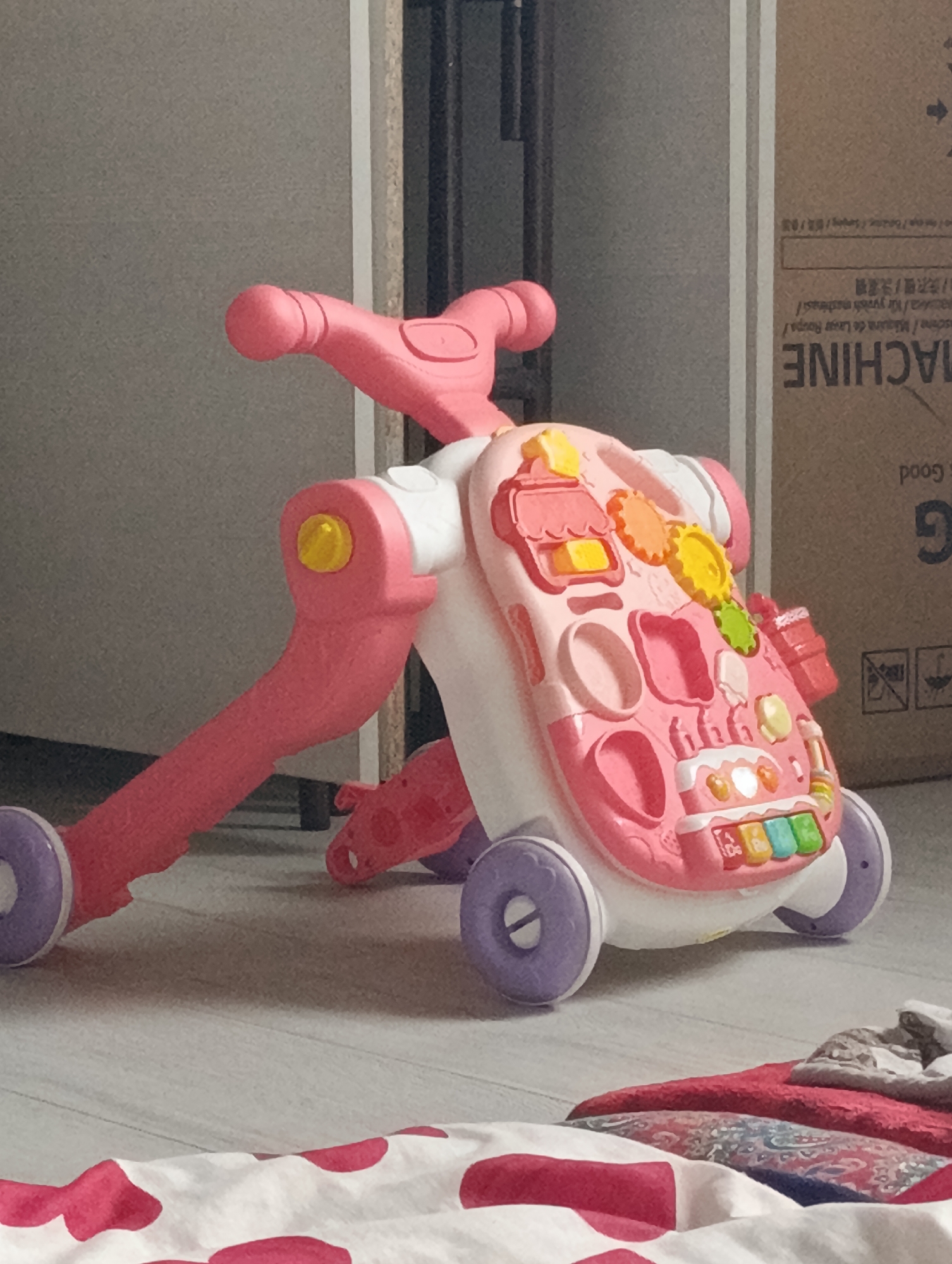 The image shows a pink and white baby walker on a light-colored floor. The walker has various colorful toys and buttons on its front panel, including what appears to be a keyboard with colorful keys and some spinning gears. The walker has four wheels, two large purple ones at the back and two smaller ones at the front. In the background, there is a grey wall and a brown cardboard box with some text and symbols on it. The floor has a wood grain pattern. A folded red and white patterned cloth is visible in the foreground. The scene is indoors and appears to be in a residential setting. No people are present in the image. The location is identified as Isaevo, Kazakhstan.
